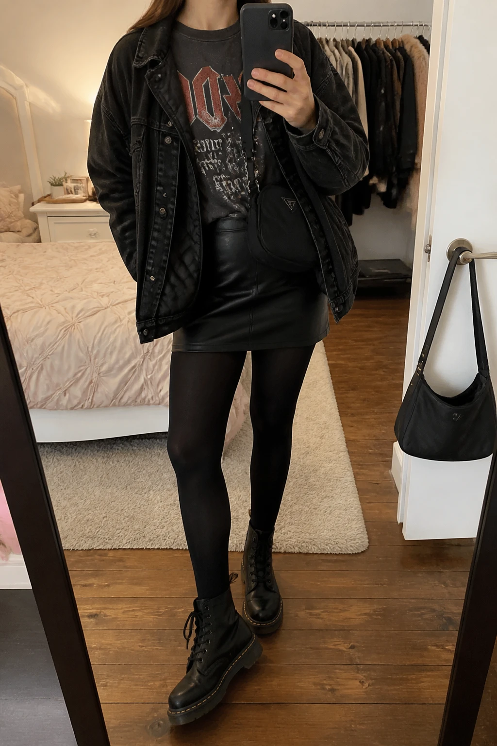Image Prompt: Mirror selfie of a woman wearing an oversized charcoal grey vintage-style band graphic cotton T-shirt tucked into a black faux-leather mini skirt, black opaque thermal tights, black leather chunky lace-up combat boots, an oversized black denim jacket with a quilted lining, and a small black nylon crossbody bag, holding a phone in front of a mirror, cropped at the NECK so the face does not show, with a relaxed casual selfie pose, in a feminine-themed bedroom, warm soft indoor lighting, a clothing rack with garments behind and a handbag hanging on a door handle, realistic worn cotton texture and natural leather folds, latest iPhone photo quality, no additional clothing or accessories.