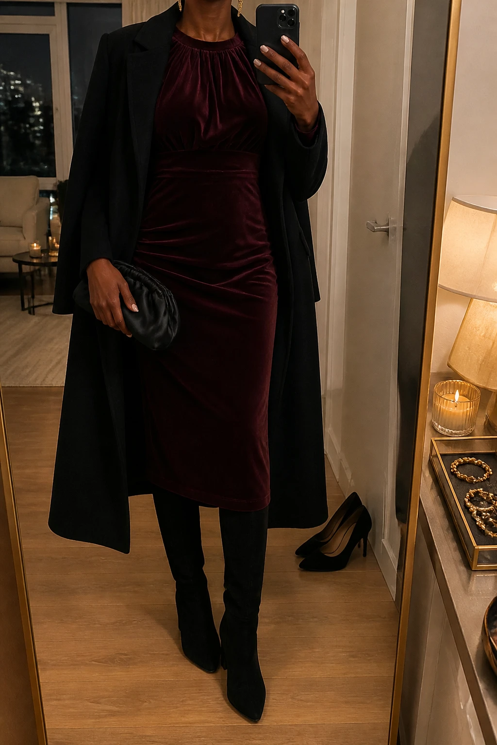 Image Prompt: Mirror selfie of a black woman wearing a burgundy long-sleeve velvet midi dress with a softly gathered neckline and fitted waist, black suede knee-high boots with low block heels, a long black tailored wool coat worn open, a small black satin evening clutch, and delicate gold drop earrings, holding a phone in front of a full-length mirror, cropped at the NECK so the face does not show, with a relaxed casual selfie pose, in a stylish city apartment, warm moody indoor lighting, a jewelry tray on the vanity and a pair of heels on the floor nearby, realistic burgundy velvet texture, natural dress folds and subtle evening shadows, latest iPhone photo quality, no additional clothing or accessories.
