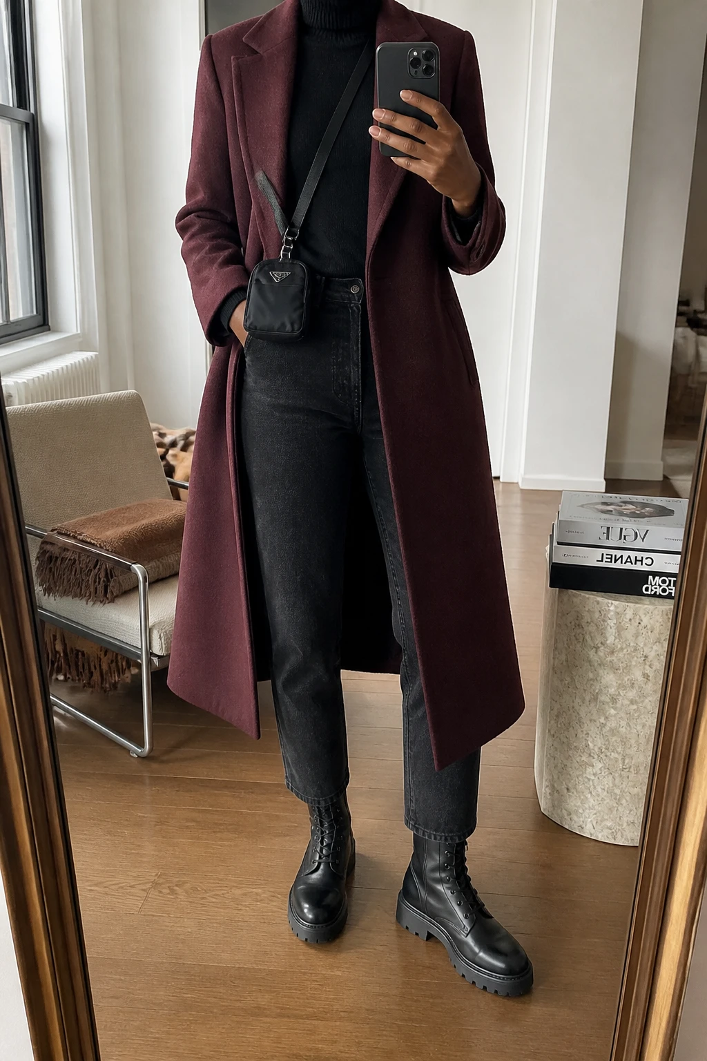 Image Prompt: Mirror selfie of a black woman wearing a long burgundy tailored wool coat over a black fitted fine-knit turtleneck sweater, dark charcoal grey straight-leg denim jeans, black leather chunky lace-up combat boots with thick rubber soles, and a small black nylon crossbody bag, holding a phone in front of a mirror, cropped at the NECK so the face does not show, with a relaxed casual selfie pose, in a stylish city apartment, soft natural window light, a stack of fashion magazines on a side table and a folded blanket on a chair, realistic burgundy wool texture, natural boot leather grain and denim creases, latest iPhone photo quality, no additional clothing or accessories.