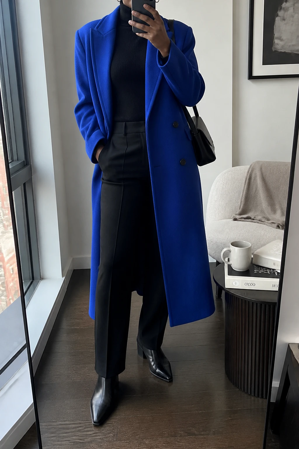 Image Prompt: Mirror selfie of a black woman wearing a long vibrant cobalt blue double-breasted tailored wool coat left open over a black fitted cashmere turtleneck sweater, black high-waisted straight-leg tailored wool trousers, black polished leather pointed-toe ankle boots with low block heels, a structured black leather shoulder bag, and small silver hoop earrings, holding a phone in front of a full-length mirror, cropped at the NECK so the face does not show, with a relaxed casual selfie pose, in a stylish city apartment, soft natural window light, a coffee mug on a side table and a folded blanket on a chair, realistic cobalt wool texture, natural black trouser creases, accurate cool blue color without oversaturation, latest iPhone photo quality, no additional clothing or accessories.
