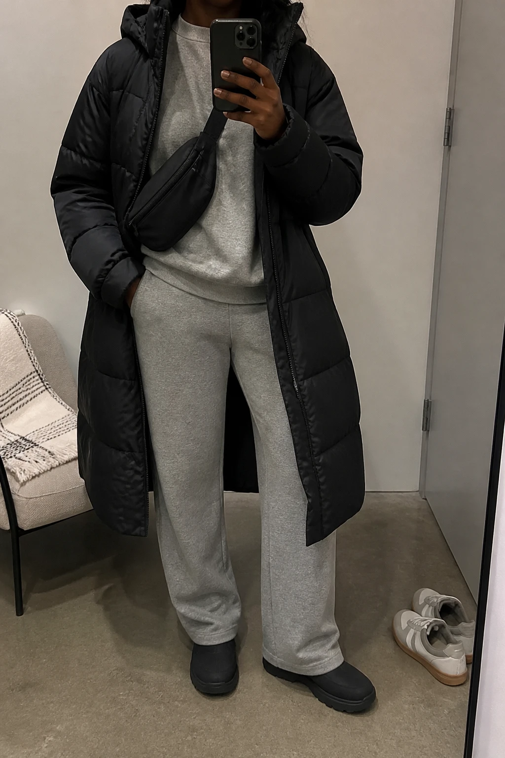 Image Prompt: Mirror selfie of a black woman wearing a long black insulated hooded puffer coat over a heather grey thick fleece-lined crewneck sweatshirt, matching heather grey straight-leg fleece-lined knit trousers, black waterproof insulated flat winter boots with rubber lug soles, a small black nylon crossbody bag, and a charcoal grey ribbed knit beanie, holding a phone in front of a mirror, cropped at the NECK so the face does not show, with a relaxed casual selfie pose, in a minimal dressing room, soft indoor lighting, a folded blanket on a chair and a pair of shoes on the floor, realistic black puffer volume, natural grey fleece folds and comfortable winter travel styling, latest iPhone photo quality, no additional clothing or accessories.
