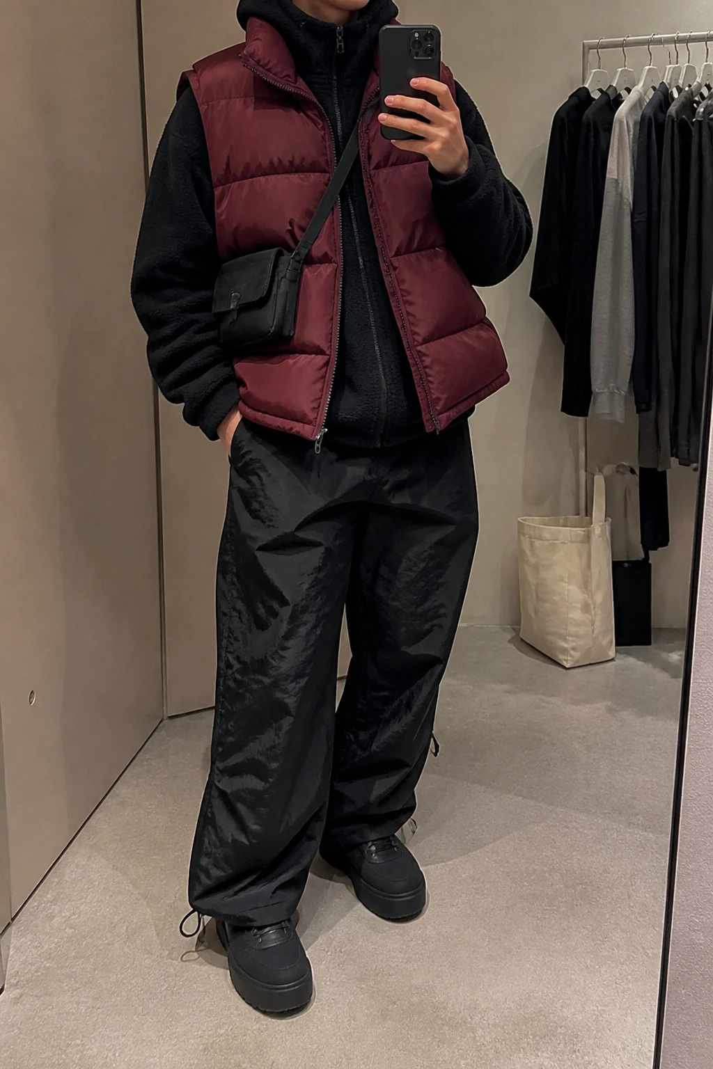 Image Prompt: Mirror selfie of a woman wearing a burgundy oversized insulated quilted puffer vest over a black heavyweight fleece-lined hoodie, black baggy nylon parachute pants with adjustable ankle cuffs, black chunky platform sneakers with thick flat soles, and a small black nylon crossbody bag, holding a phone in front of a mirror, cropped at the NECK so the face does not show, with a relaxed casual selfie pose, in a minimal dressing room, soft indoor lighting, a tote bag resting on the floor and a clothing rack with garments behind, realistic burgundy quilted fabric, natural black parachute pant folds and casual streetwear styling, latest iPhone photo quality, no additional clothing or accessories.