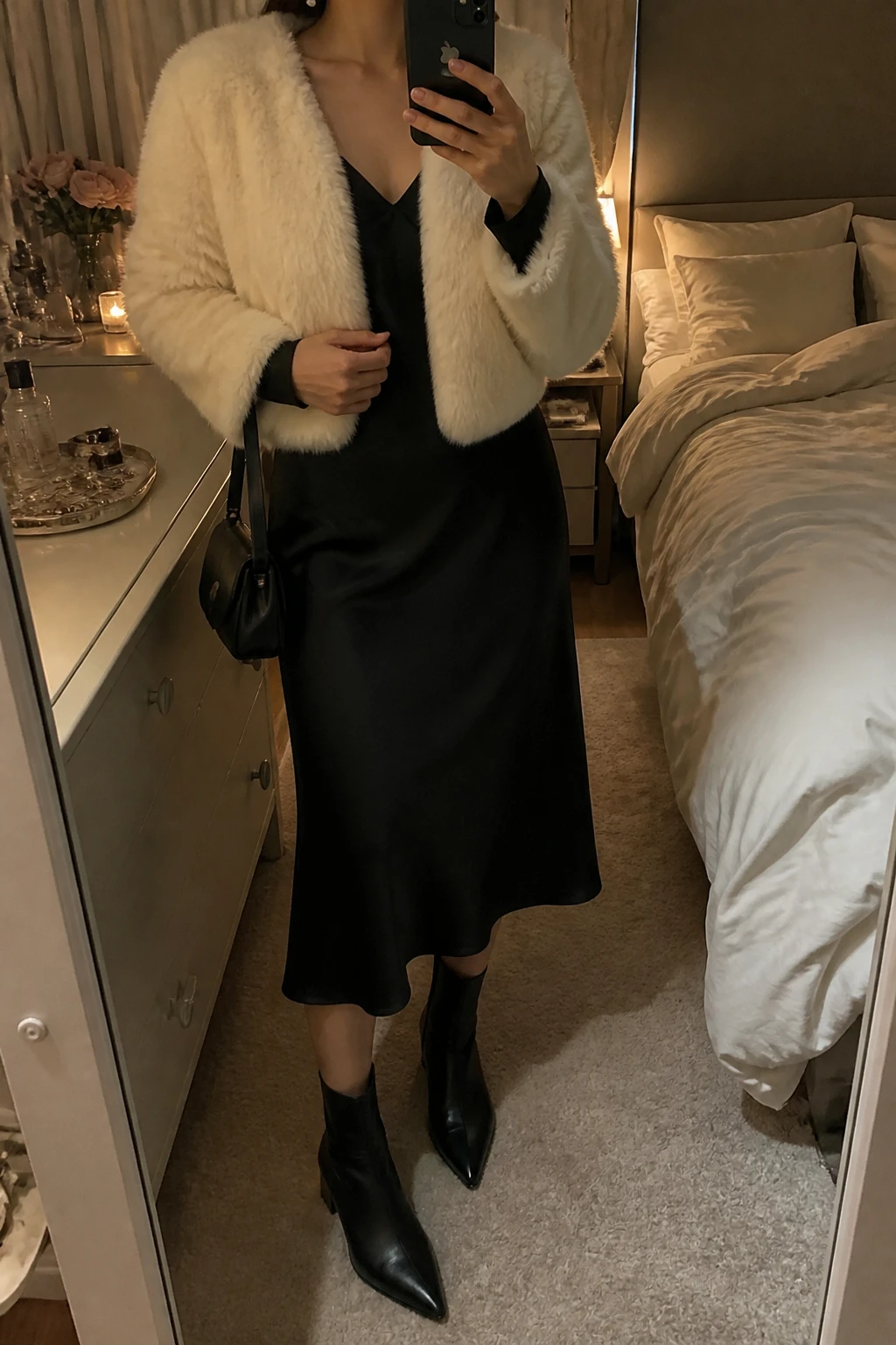 Image Prompt: Mirror selfie of a woman wearing a cropped cream faux-fur jacket over a black long-sleeve satin midi dress with a modest V-neckline and softly fitted waist, black leather pointed-toe ankle boots with low block heels, a small black satin shoulder bag, and simple pearl stud earrings, holding a phone in front of a mirror, cropped at the NECK so the face does not show, with a relaxed casual selfie pose, in a feminine-themed bedroom, warm moody lighting, soft pillows on an unmade bed and a jewelry tray on the vanity, realistic cream faux-fur texture, natural black satin folds and subtle evening atmosphere, latest iPhone photo quality, no additional clothing or accessories.