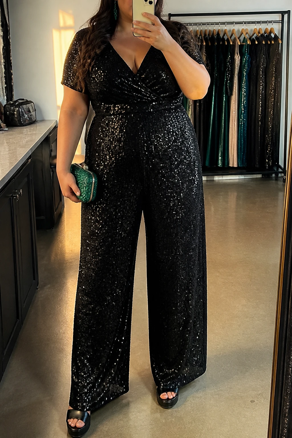 Mirror selfie of a plus size woman wearing a black sequin wide-leg jumpsuit with short sleeves, a V-neckline, and a softly defined waist, black platform sandals, a small emerald green crystal-covered clutch, and emerald drop earrings, clearly showing the exact jumpsuit shape, black sequins, green accessories, colors, and footwear listed, holding a phone in front of a mirror, cropped at the neck so the face is hidden, relaxed glamorous selfie pose, in a fashion studio mirror corner with evening golden-hour lighting, a clothing rack with garments behind her and a cosmetic bag on the counter, realistic formal birthday appearance, latest iPhone photo quality.