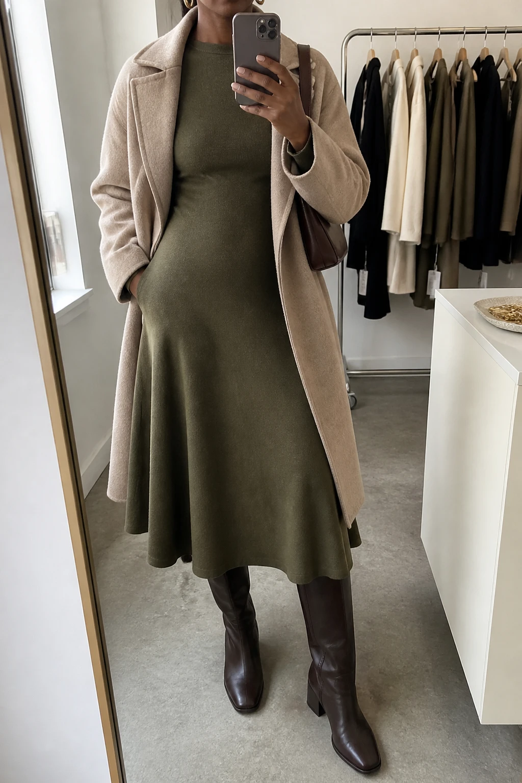 Image Prompt: Mirror selfie of a black woman wearing an olive green long-sleeve fine-knit midi dress with a softly flared skirt, dark brown leather knee-high low block-heel boots, a warm beige wool wrap coat worn open, a small dark brown leather shoulder bag, and minimal gold oval earrings, holding a phone in front of a mirror, cropped at the NECK so the face does not show, with a relaxed casual selfie pose, in a fashion studio mirror corner, soft natural window light, a clothing rack with garments behind and a jewelry tray on the vanity, realistic olive knit fabric, natural skirt folds and warm-neutral color rendering, latest iPhone photo quality, no additional clothing or accessories.