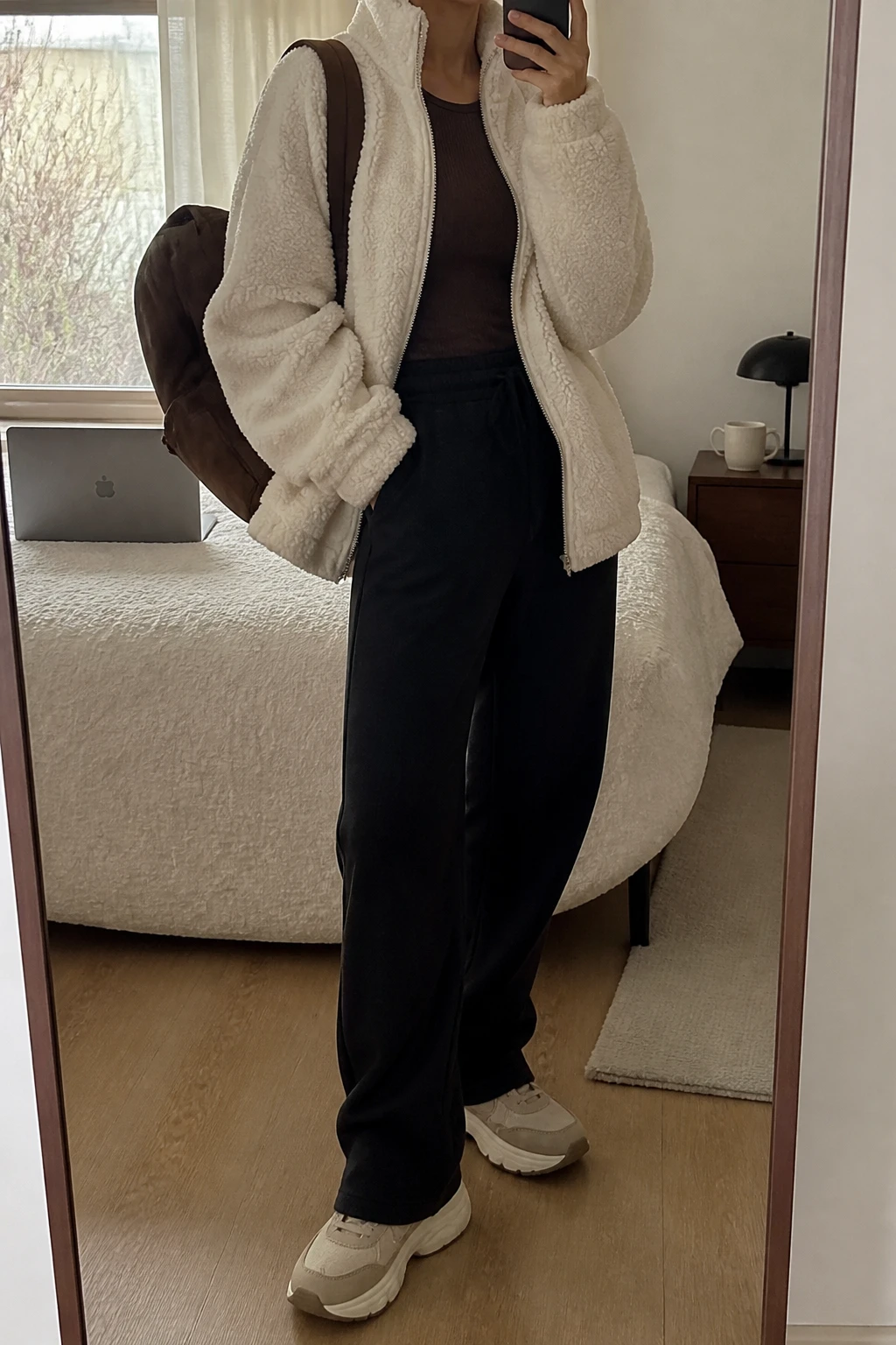 Image Prompt: Mirror selfie of a woman wearing a cream oversized teddy-fleece zip-front jacket over a chocolate brown fitted ribbed long-sleeve top, black high-waisted straight-leg fleece sweatpants, beige chunky cushioned sneakers with cream rubber soles, and a medium chocolate brown nylon backpack, holding a phone in front of a mirror, cropped at the NECK so the face does not show, with a relaxed casual selfie pose, in a cozy bedroom mirror setup, soft natural window light, a laptop on the bed and a coffee mug on a bedside table, realistic cream teddy-fleece texture, natural black sweatpant folds and subdued warm-neutral colors, latest iPhone photo quality, no additional clothing or accessories.