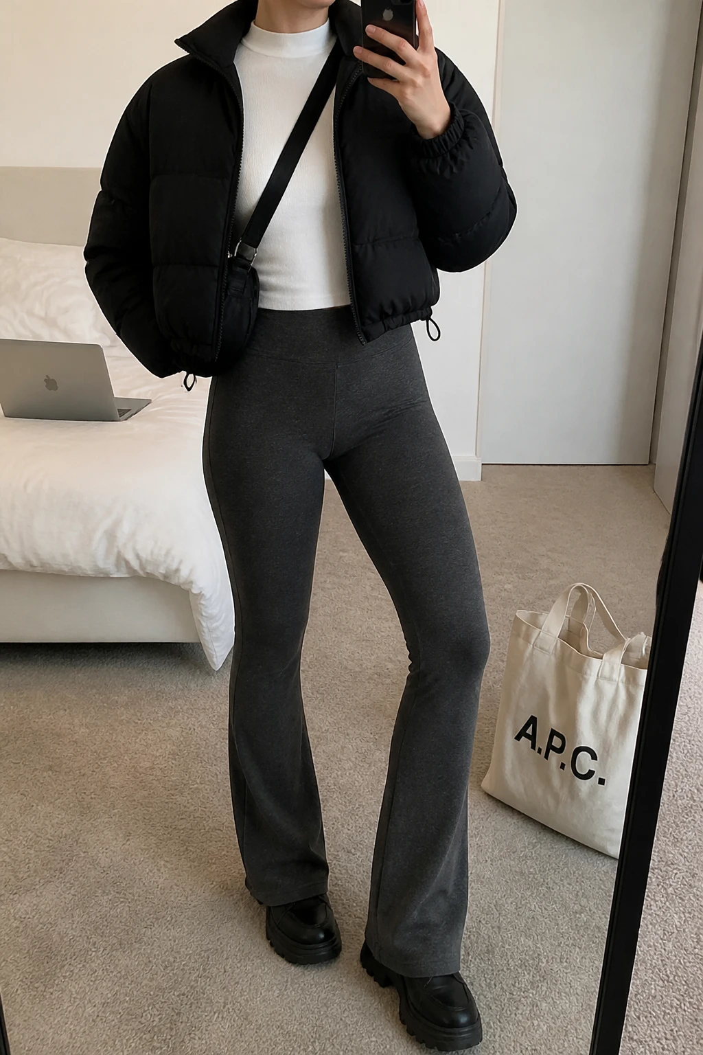 Image Prompt: Mirror selfie of a woman wearing a black cropped insulated quilted puffer jacket over a white fitted long-sleeve thermal mock-neck top, charcoal grey high-waisted fleece-lined flared leggings, black chunky leather sneakers with thick rubber soles, and a small black nylon crossbody bag, holding a phone in front of a mirror, cropped at the NECK so the face does not show, with a relaxed casual selfie pose, in a cozy bedroom mirror setup, soft indoor lighting, a tote bag resting on the floor and a laptop on the bed, realistic black puffer volume, natural charcoal grey stretch fabric and casual everyday proportions, latest iPhone photo quality, no additional clothing or accessories.