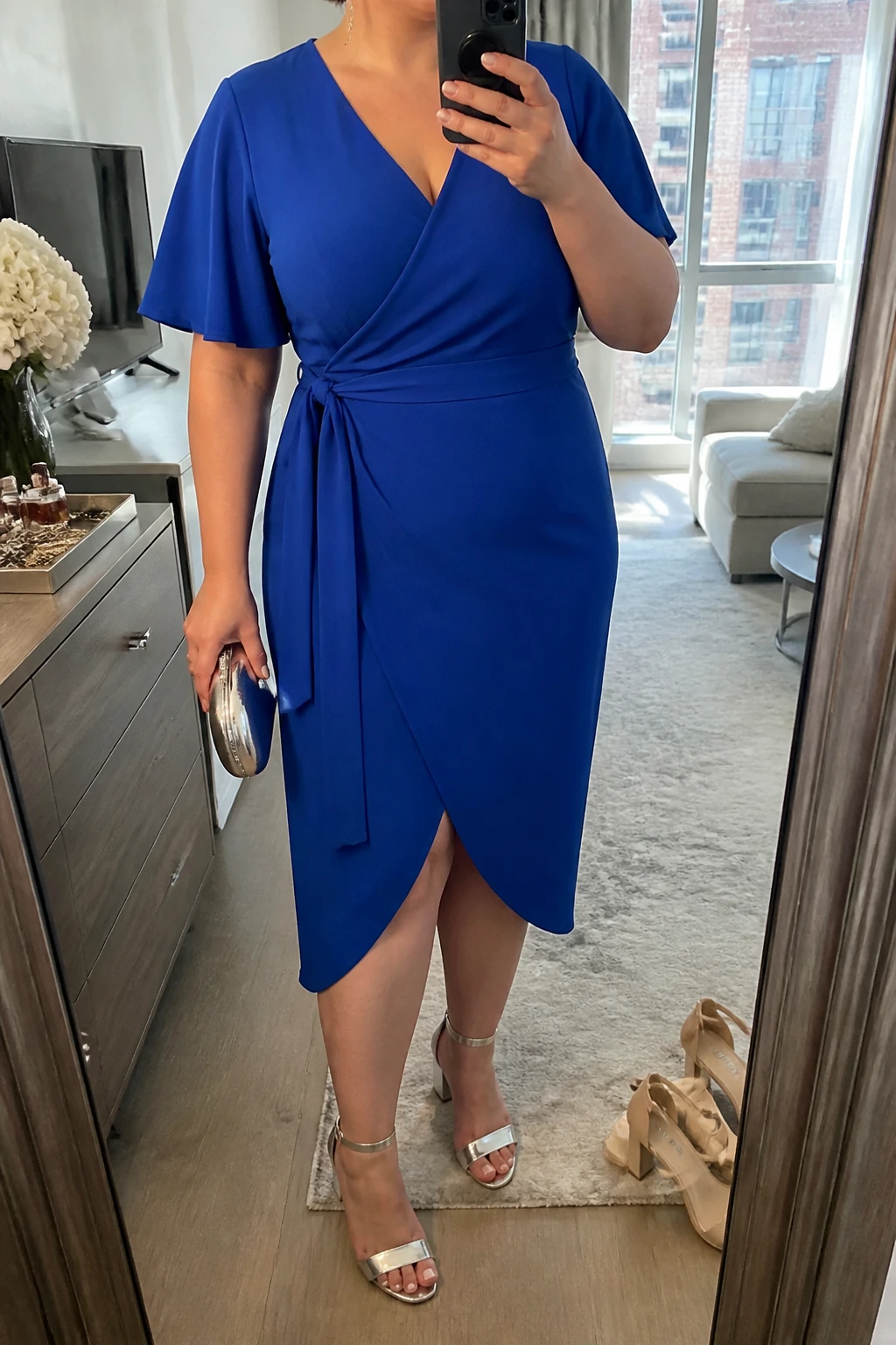 Mirror selfie of a plus size woman wearing a cobalt blue crepe wrap midi dress with short flutter sleeves, a V-neckline, and a matching waist tie, metallic silver block-heel sandals, a small silver structured clutch, and delicate silver drop earrings, clearly showing the exact clothing items, cobalt color, crepe fabric, wrap shape, footwear, and accessories listed, holding a phone in front of a mirror, cropped at the neck so the face does not show, relaxed confident selfie pose, in a stylish city apartment with natural window light, a jewelry tray on the dresser and a pair of heels on the floor nearby, natural unstaged graduation-day appearance, latest iPhone photo quality.