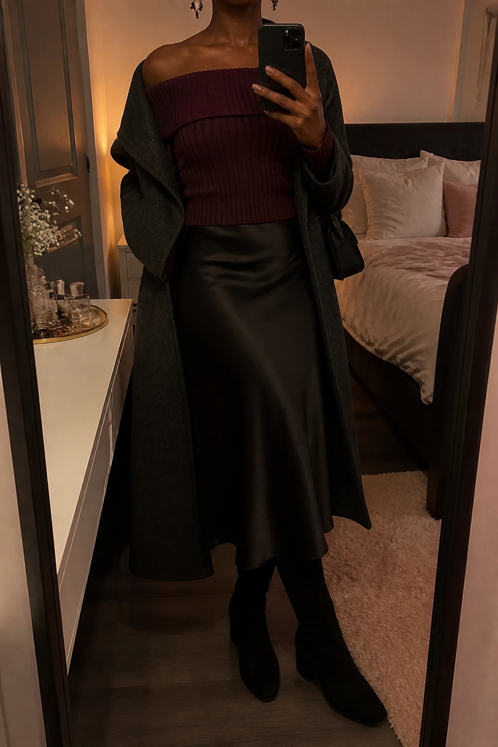 Image Prompt: Mirror selfie of a black woman wearing a deep burgundy fitted off-shoulder ribbed knit sweater with a folded neckline tucked into a black satin bias-cut midi skirt, black suede knee-high boots with low block heels, a long charcoal grey tailored wool coat worn open, a small black satin shoulder bag, and delicate silver drop earrings, holding a phone in front of a full-length mirror, cropped at the NECK so the face does not show, with a relaxed casual selfie pose, in a feminine-themed bedroom, warm moody lighting, soft pillows on an unmade bed and a jewelry tray on the vanity, realistic burgundy ribbed knit texture, natural black satin drape, soft evening shadows, latest iPhone photo quality, no additional clothing or accessories.