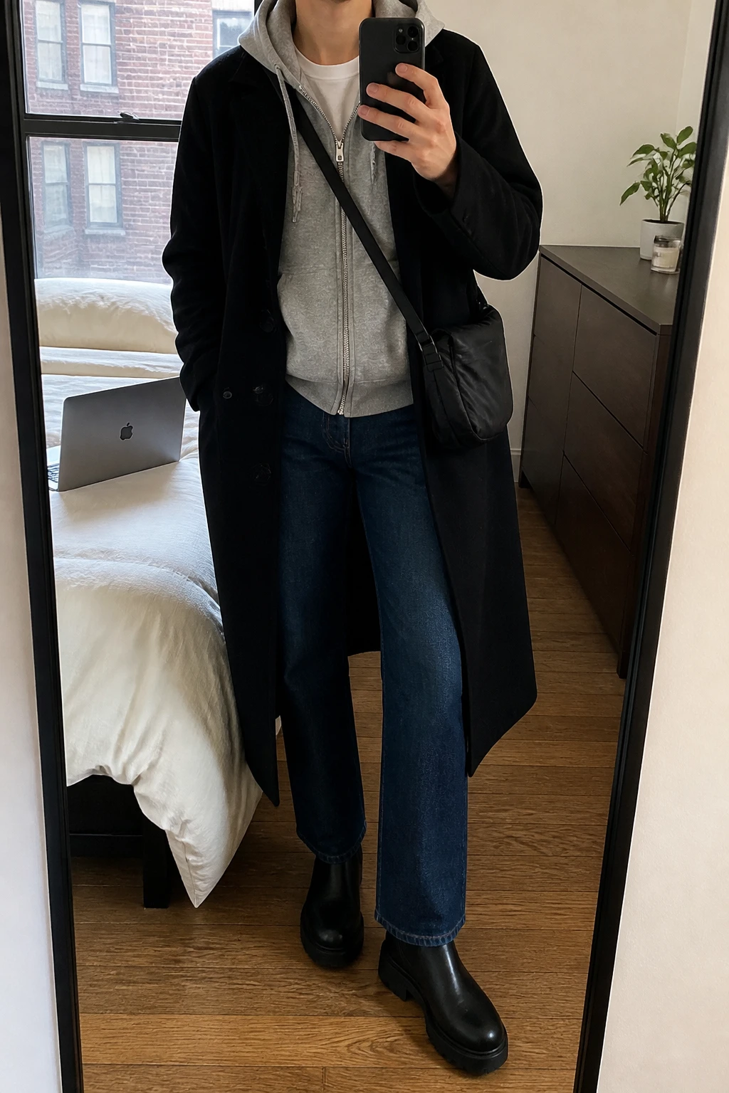 Image Prompt: Mirror selfie of a woman wearing a long black single-breasted wool coat open over a heather grey thick fleece zip-up hoodie, a white cotton crewneck T-shirt visible beneath the partly unzipped hoodie, dark blue straight-leg jeans, black leather chunky Chelsea boots, and a black nylon crossbody bag, holding a phone in front of a mirror, cropped at the NECK so the face does not show, with a relaxed casual selfie pose, in a stylish city apartment, natural window light, a laptop on the bed and a small plant on a dresser, realistic hoodie folds and wool coat drape, latest iPhone photo quality, no additional clothing or accessories.