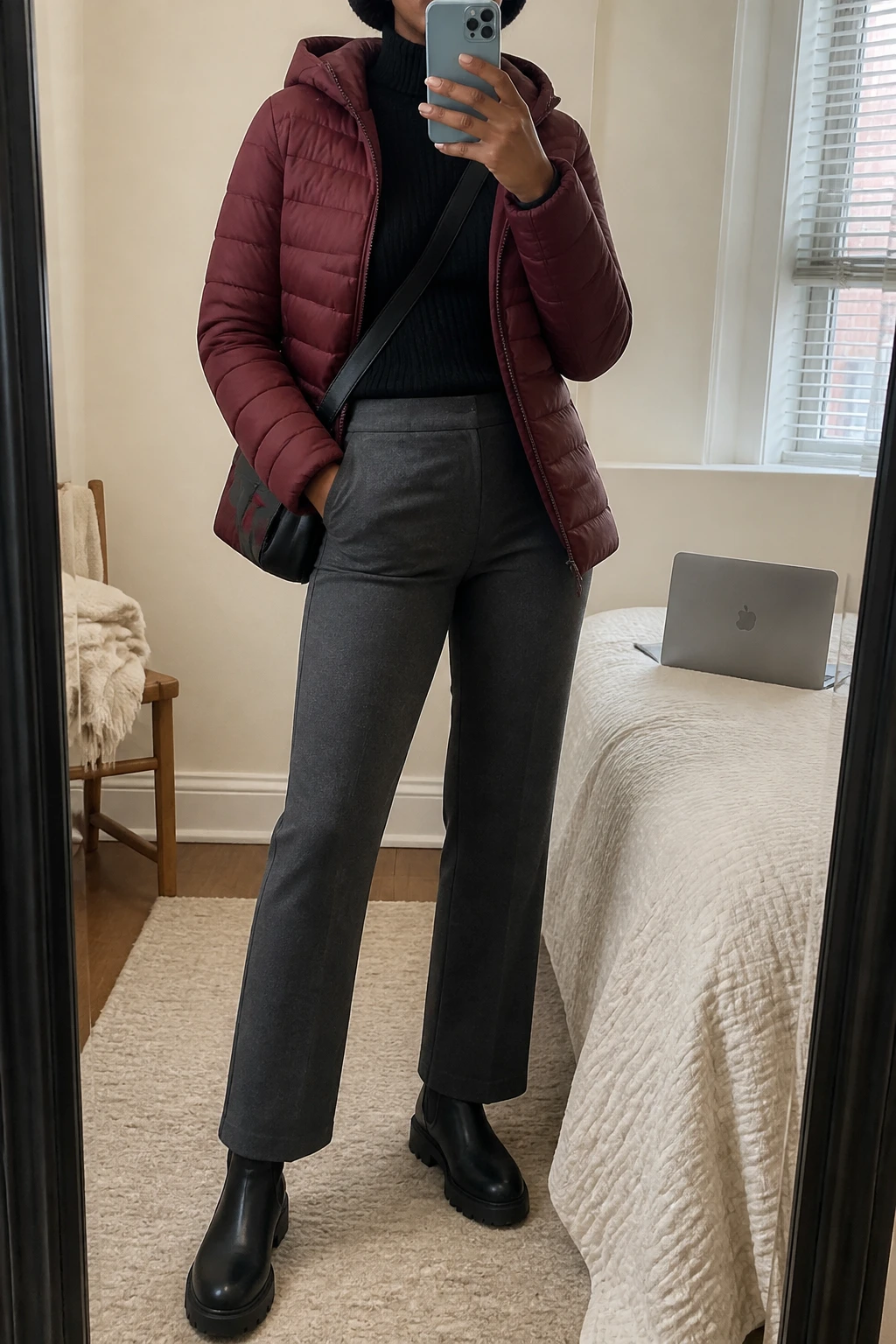 Image Prompt: Mirror selfie of a black woman wearing a burgundy insulated hooded hip-length puffer jacket over a black fitted thermal mock-neck sweater, charcoal grey high-waisted straight-leg thick ponte trousers, black waterproof leather chunky flat Chelsea boots with grippy rubber soles, a small black nylon crossbody bag, and a black ribbed knit beanie, holding a phone in front of a mirror, cropped at the NECK so the face does not show, with a relaxed casual selfie pose, in a cozy bedroom mirror setup, soft natural window light, a laptop on the bed and a folded blanket on a chair, realistic burgundy quilted fabric, natural grey ponte folds and warm black thermal fabric, latest iPhone photo quality, no additional clothing or accessories.