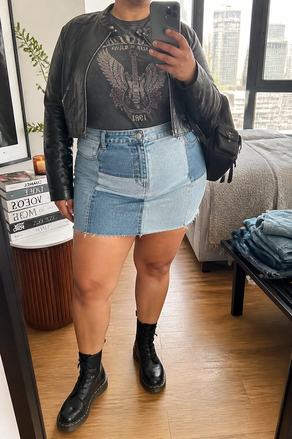 Mirror selfie of a plus size woman wearing a blue patchwork denim mini skirt with mixed light and medium-wash panels, a fitted faded black graphic T-shirt tucked into the skirt, a cropped black leather jacket, black lace-up combat boots, and a small black nylon backpack, clearly showing the exact clothing items, denim panels, graphic print, black jacket, footwear, and accessories listed, holding a phone in front of a mirror, cropped at the neck so her face is hidden, relaxed edgy selfie stance, in a stylish city apartment with natural window light, a stack of fashion magazines on a side table and folded denim jeans on a bench, realistic casual concert styling, latest iPhone photo quality.