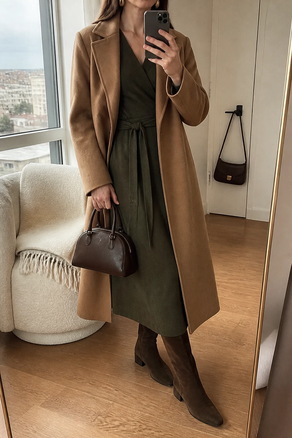 Image Prompt: Mirror selfie of a woman wearing an olive green long-sleeve wool-blend wrap midi dress with a modest V-neckline and softly tied waist, chocolate brown suede knee-high boots with low block heels, a long camel tailored wool coat worn open, a structured dark brown leather handbag, and small gold oval earrings, holding a phone in front of a mirror, cropped at the NECK so the face does not show, with a relaxed casual selfie pose, in a stylish city apartment, soft natural window light, a folded blanket on a chair and a handbag hanging on a door handle, realistic olive wool dress texture, natural suede boots and warm neutral styling, latest iPhone photo quality, no additional clothing or accessories.