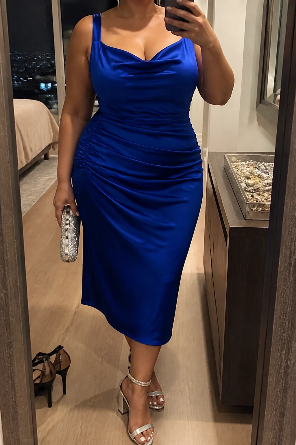Mirror selfie of a plus size woman wearing a cobalt blue satin midi dress with wide supportive straps, a softly draped neckline, and gentle ruching along one side of the waist, metallic silver block heels with crystal ankle straps, a small silver rhinestone clutch, and silver drop earrings, clearly showing the exact clothing items, cobalt satin, ruching, colors, footwear, and accessories listed, holding a phone in front of a mirror, cropped at the neck so the face does not show, relaxed confident selfie pose, in a stylish city apartment with warm moody lighting, a jewelry tray on the dresser and a pair of heels on the floor nearby, natural unstaged birthday dinner appearance, latest iPhone photo quality.