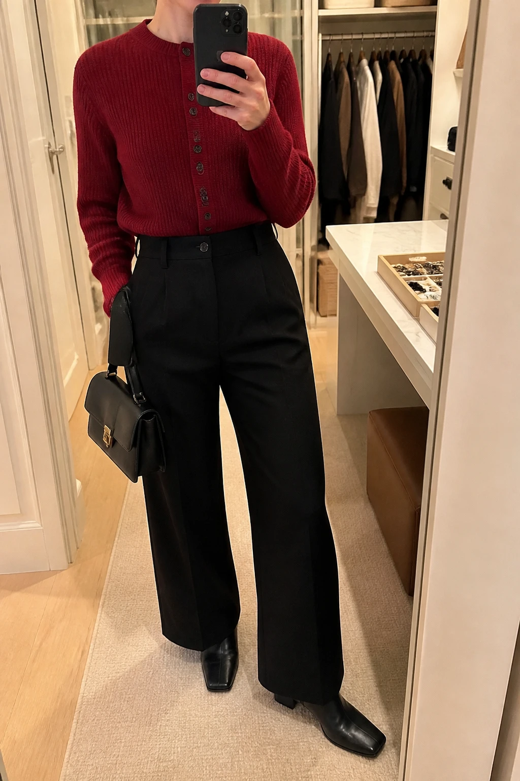 Image Prompt: Mirror selfie of a woman wearing a cherry red fitted chunky-knit button-front cardigan fully buttoned and tucked into black high-waisted wide-leg wool trousers, black leather square-toe ankle boots, a small black leather top-handle bag, and small gold dome earrings, holding a phone in front of a mirror, cropped at the NECK so the face does not show, with a relaxed casual selfie pose, in a walk-in closet mirror, warm soft indoor lighting, an open closet with clothes visible and a jewelry tray on the vanity, realistic cherry red color and fabric texture, latest iPhone photo quality, no additional clothing or accessories.
