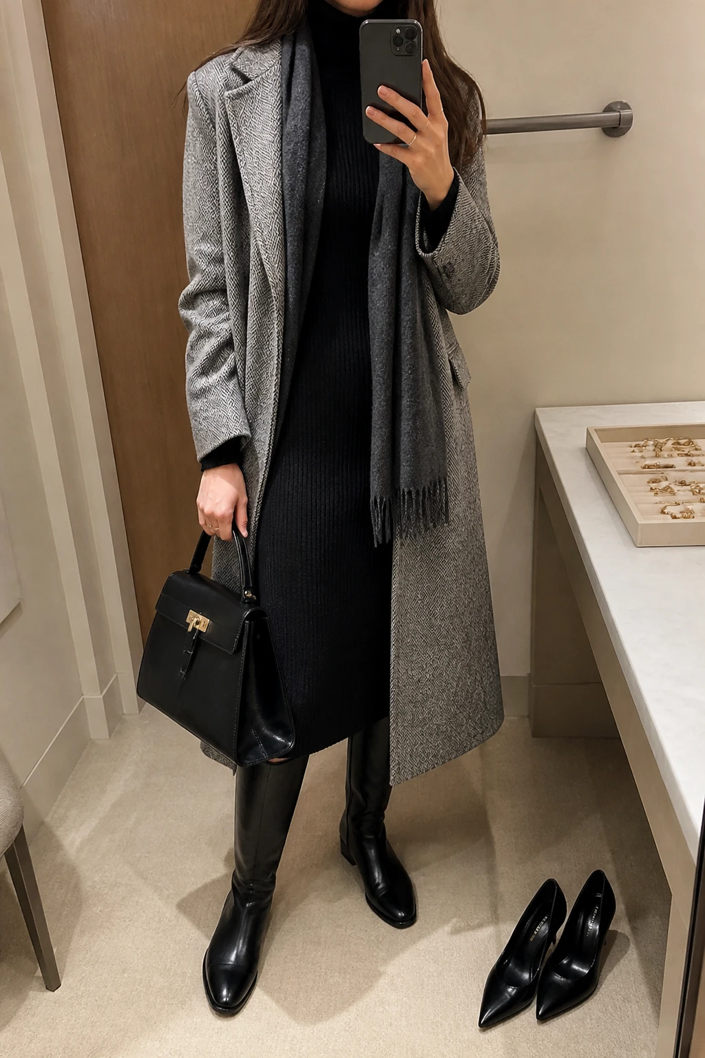 Image Prompt: Mirror selfie of a woman wearing a long grey herringbone wool coat over a black long-sleeve ribbed knit midi turtleneck dress, black fleece-lined opaque tights, black leather knee-high flat boots with rubber soles, a structured black leather top-handle bag, and a charcoal grey wool scarf, holding a phone in front of a mirror, cropped at the NECK so the face does not show, with a relaxed casual selfie pose, in a minimal dressing room, soft indoor lighting, a jewelry tray on the vanity and a pair of heels on the floor nearby, natural herringbone texture and realistic dress proportions, latest iPhone photo quality, no additional clothing or accessories.