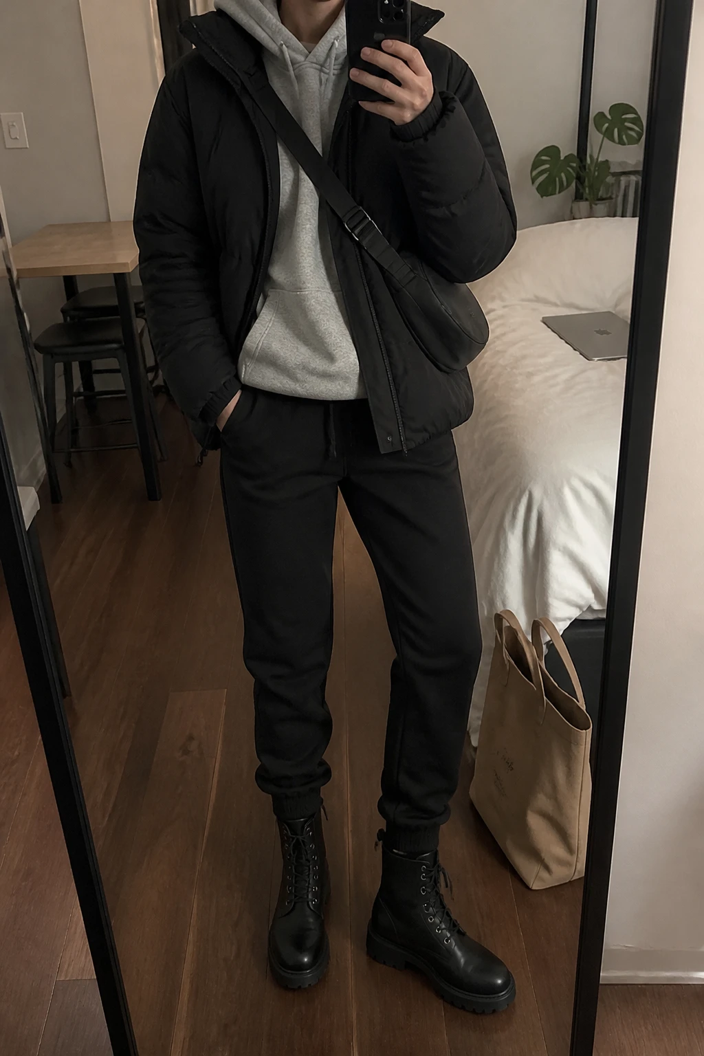 Image Prompt: Mirror selfie of a woman wearing a black oversized insulated quilted puffer jacket over a heather grey thick fleece-lined hoodie, black straight-leg thermal jogger pants, black waterproof leather lace-up ankle boots with flat lug soles, and a small black nylon crossbody bag, holding a phone in front of a mirror, cropped at the NECK so the face does not show, with a relaxed casual selfie pose, in a stylish city apartment, soft indoor lighting, a tote bag resting on the floor and a laptop on the bed, natural puffer volume, realistic fleece texture and casual winter proportions, latest iPhone photo quality, no additional clothing or accessories.