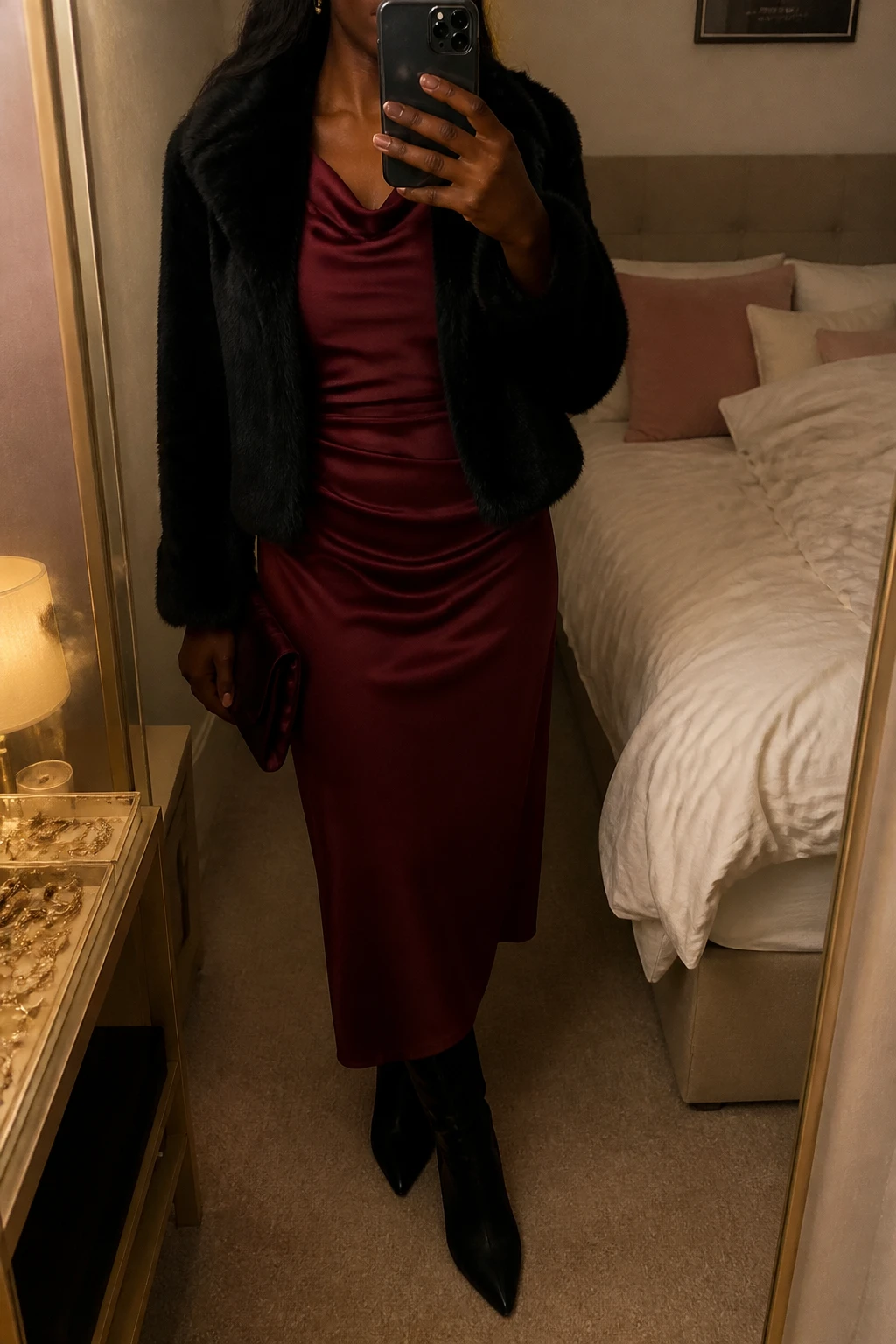 Image Prompt: Mirror selfie of a black woman wearing a burgundy long-sleeve satin midi dress with a softly draped neckline and fitted waist, a cropped black faux-fur jacket worn open, black suede pointed-toe heeled ankle boots, a small burgundy satin clutch, and delicate gold stud earrings, holding a phone in front of a mirror, cropped at the NECK so the face does not show, with a relaxed casual selfie pose, in a feminine-themed bedroom, warm moody lighting, soft pillows on an unmade bed and a jewelry tray on the vanity, realistic burgundy satin draping, natural black faux-fur texture, subtle warm evening shadows, latest iPhone photo quality, no additional clothing or accessories.
