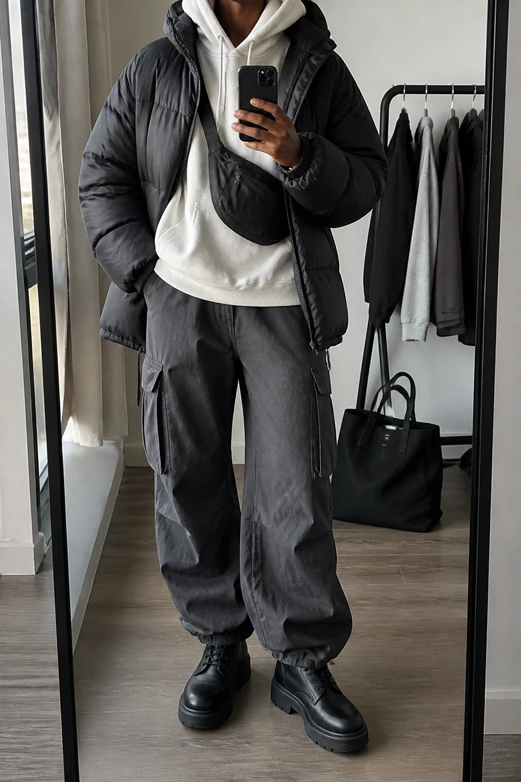 Image Prompt: Mirror selfie of a black woman wearing a black oversized insulated quilted puffer jacket left open over a white heavyweight fleece-lined hoodie, charcoal grey baggy cargo trousers with large side pockets, black leather chunky lace-up boots with thick flat rubber soles, and a small black nylon crossbody bag worn across the chest, holding a phone in front of a full-length mirror, cropped at the NECK so the face does not show, with a relaxed casual selfie pose, in a stylish city apartment, soft natural window light, a tote bag resting on the floor and a clothing rack with garments behind, realistic puffer volume, natural cargo fabric creases, casual everyday streetwear styling, latest iPhone photo quality, no additional clothing or accessories.