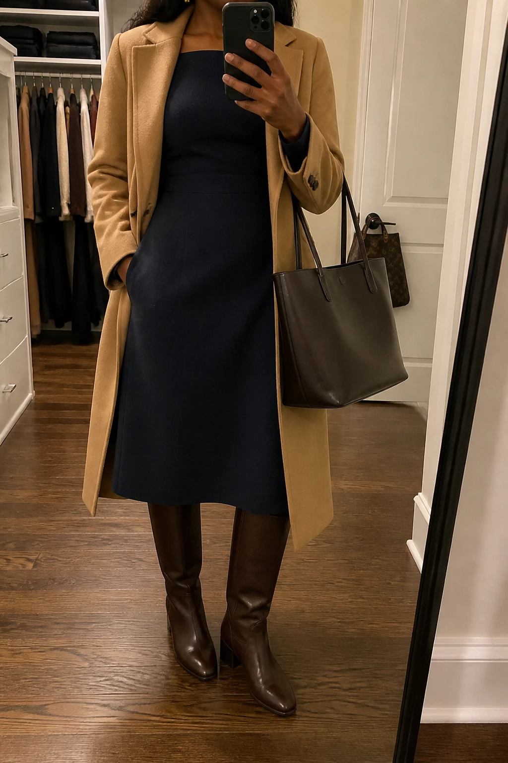 Image Prompt: Mirror selfie of a black woman wearing a navy long-sleeve wool-blend midi dress with a modest square neckline and fitted waist, chocolate brown leather knee-high low block-heel boots, a camel long tailored wool coat worn open, a structured dark brown leather work tote, and small gold stud earrings, holding a phone in front of a mirror, cropped at the NECK so the face does not show, with a relaxed casual selfie pose, in a walk-in closet mirror, warm soft indoor lighting, an open closet with clothes visible and a handbag hanging on a door handle, natural wool dress folds and realistic leather texture, latest iPhone photo quality, no additional clothing or accessories.