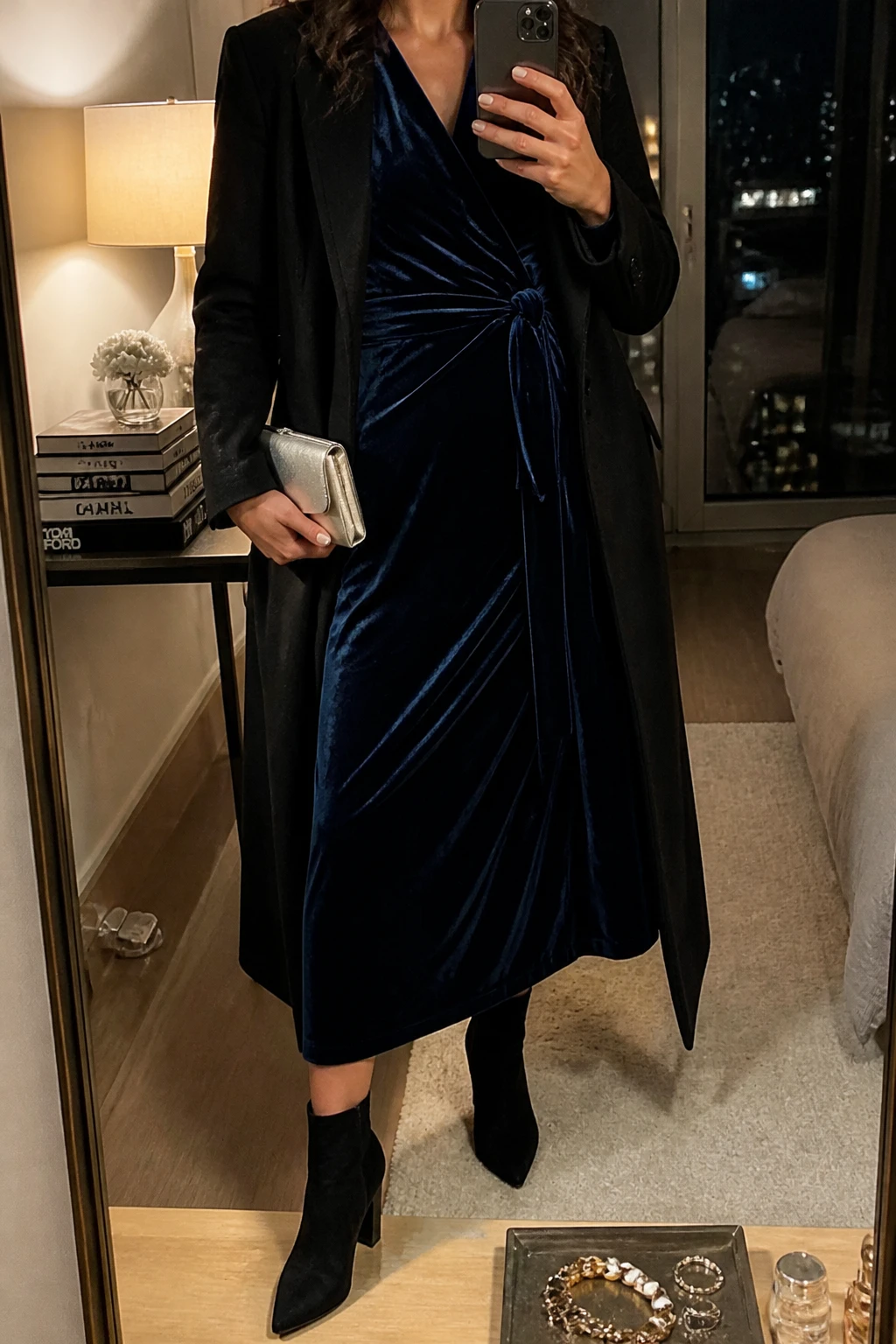 Image Prompt: Mirror selfie of a woman wearing a midnight navy long-sleeve velvet wrap midi dress with a softly tied waist, black suede pointed-toe ankle boots with block heels, a long black tailored wool coat worn open, a small silver satin evening clutch, and silver crystal drop earrings, holding a phone in front of a mirror, cropped at the NECK so the face does not show, with a relaxed casual selfie pose, in a stylish city apartment, warm moody indoor lighting, a stack of fashion magazines on a side table and a jewelry tray on the vanity, natural navy velvet reflections, realistic dress draping and subtle warm shadows, latest iPhone photo quality, no additional clothing or accessories.