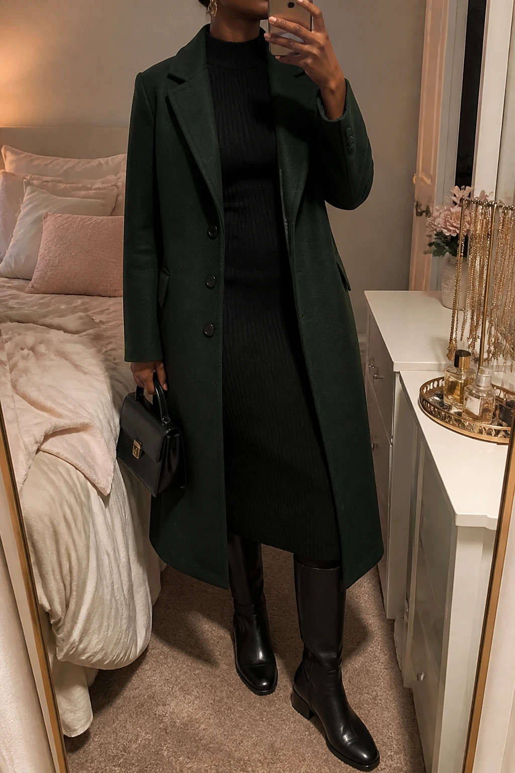 Image Prompt: Mirror selfie of a black woman wearing a long forest green single-breasted tailored wool coat over a black long-sleeve ribbed knit midi dress with a high neckline and gently fitted waist, black leather knee-high boots with low block heels, a small structured black leather handbag, and delicate gold oval earrings, holding a phone in front of a mirror, cropped at the NECK so the face does not show, with a relaxed casual selfie pose, in a feminine-themed bedroom, warm soft indoor lighting, soft pillows on an unmade bed and a jewelry tray on the vanity, realistic forest green wool texture, natural black ribbed knit folds and soft evening shadows, latest iPhone photo quality, no additional clothing or accessories.