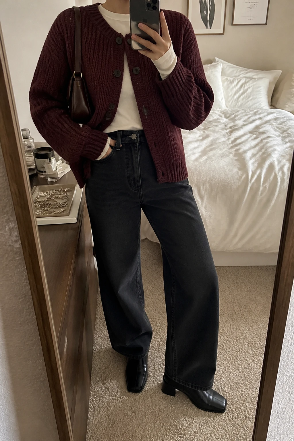 Image Prompt: Mirror selfie of a woman wearing a burgundy chunky-knit button-front cardigan over an ivory fitted cotton long-sleeve crewneck top, washed black high-waisted wide-leg denim jeans, black leather square-toe ankle boots with low block heels, and a small chocolate brown leather shoulder bag, holding a phone in front of a mirror, cropped at the NECK so the face does not show, with a relaxed casual selfie pose, in a cozy bedroom mirror setup, warm soft indoor lighting, soft pillows on an unmade bed and a jewelry tray on the vanity, realistic burgundy knit texture, natural black denim folds and comfortable casual proportions, latest iPhone photo quality, no additional clothing or accessories.