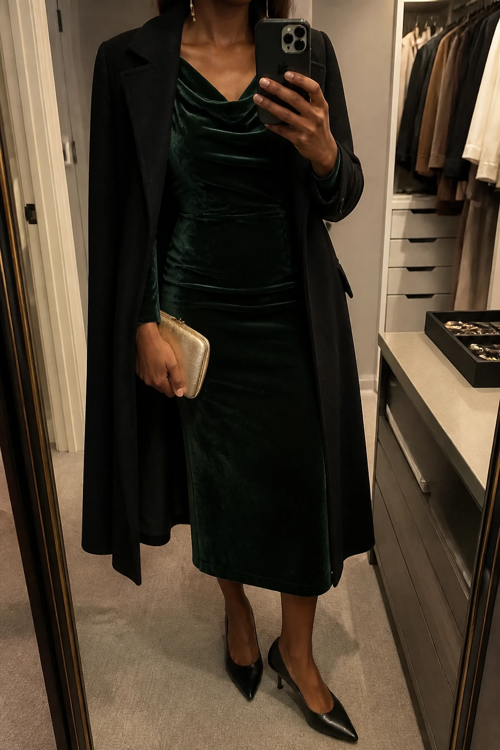 Image Prompt: Mirror selfie of a black woman wearing a forest green long-sleeve velvet midi dress with a gently draped neckline and fitted waist, black leather pointed-toe kitten heels, a small champagne gold satin clutch, delicate gold drop earrings, and a long black tailored wool coat worn open, holding a phone in front of a mirror, cropped at the NECK so the face does not show, with a relaxed casual selfie pose, in a walk-in closet mirror, warm moody indoor lighting, an open closet with clothes visible and a jewelry tray on the vanity, realistic green velvet fabric, subdued gold accessories and natural elegant posture, latest iPhone photo quality, no additional clothing or accessories.