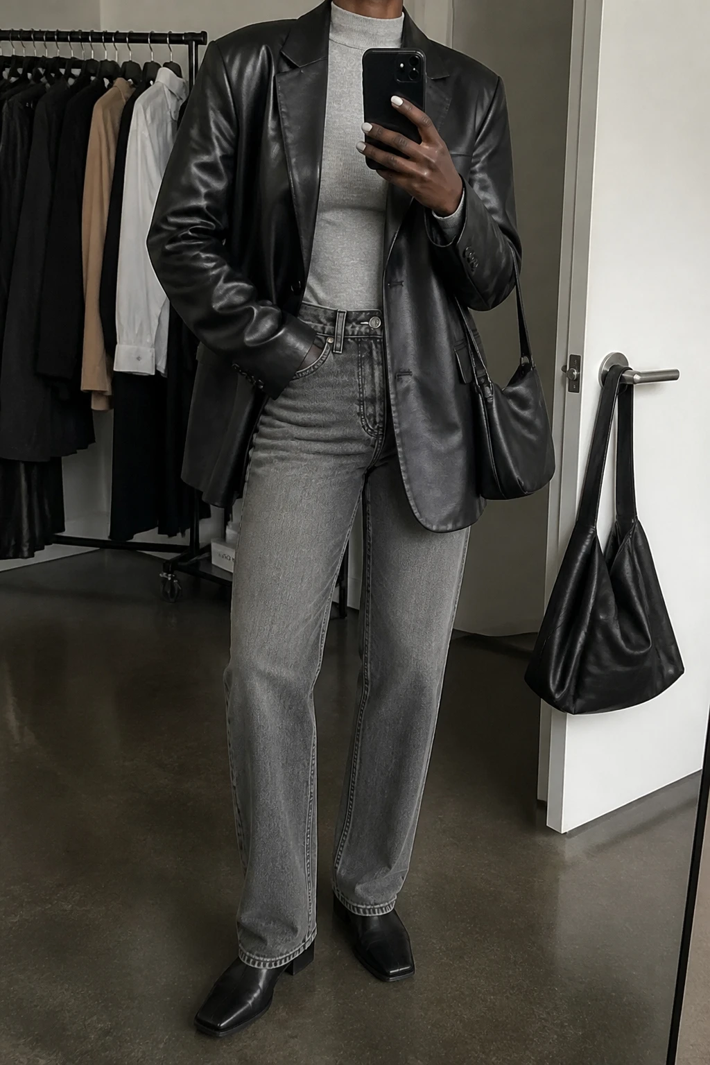 Image Prompt: Mirror selfie of a black woman wearing an oversized black faux-leather single-breasted blazer over a heather grey fitted cashmere mock-neck sweater, washed grey high-waisted straight-leg denim jeans, black leather square-toe ankle boots with flat soles, and a small black leather shoulder bag, holding a phone in front of a mirror, cropped at the NECK so the face does not show, with a relaxed casual selfie pose, in a fashion studio mirror corner, soft indoor lighting, a clothing rack with garments behind and a handbag hanging on a door handle, realistic black leather blazer grain, natural heather grey knit texture and grey denim creases, latest iPhone photo quality, no additional clothing or accessories.
