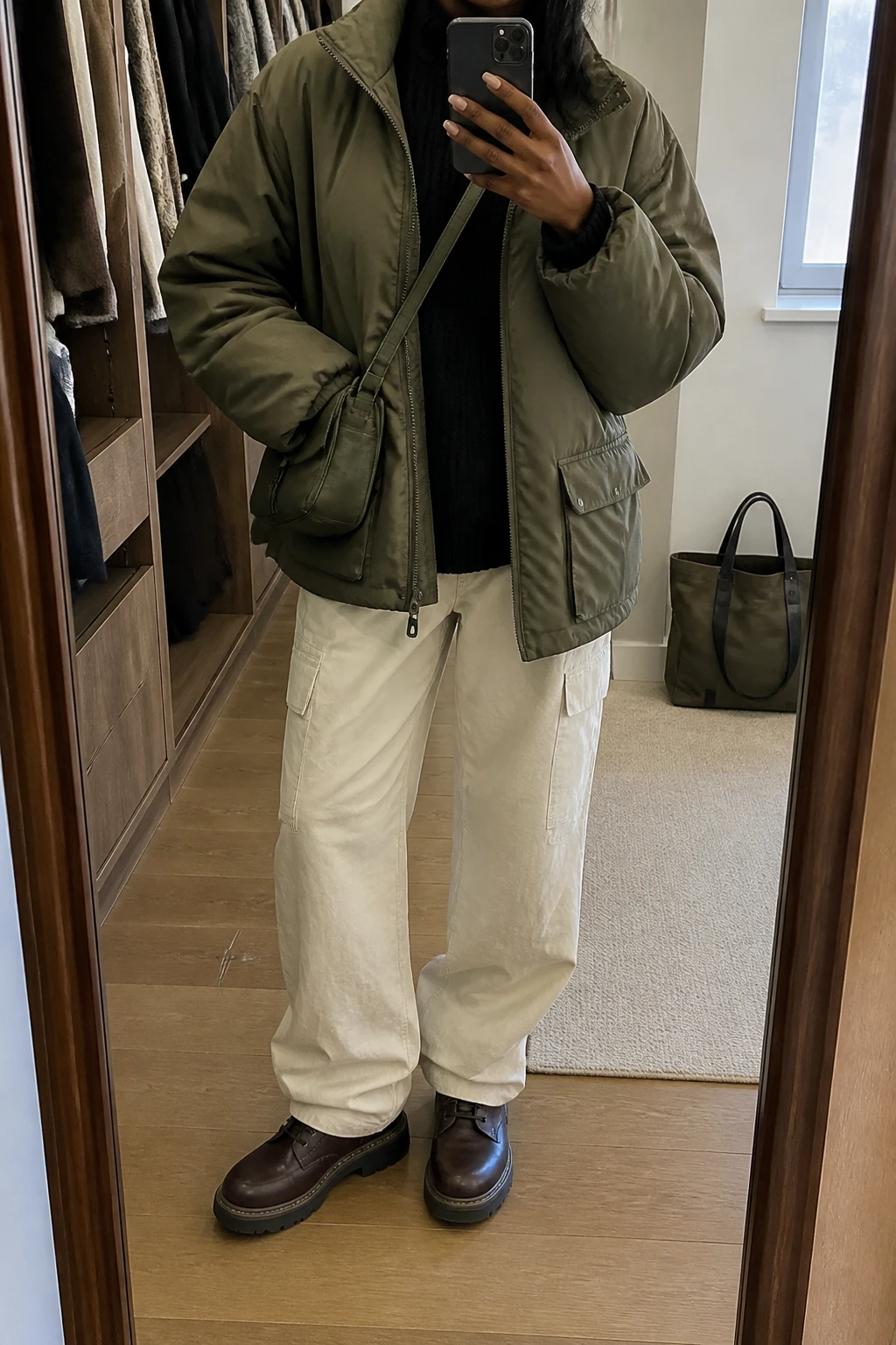 Image Prompt: Mirror selfie of a black woman wearing an oversized olive green insulated utility jacket with large front pockets over a black heavyweight ribbed mock-neck sweater, cream baggy straight-leg cargo trousers, dark brown leather chunky lace-up boots with rubber soles, and a small dark olive nylon crossbody bag, holding a phone in front of a mirror, cropped at the NECK so the face does not show, with a relaxed casual selfie pose, in a walk-in closet mirror, soft natural window light, an open closet with clothes visible and a tote bag resting on the floor, realistic olive cotton fabric, natural cream cargo trouser folds and practical winter streetwear styling, latest iPhone photo quality, no additional clothing or accessories.