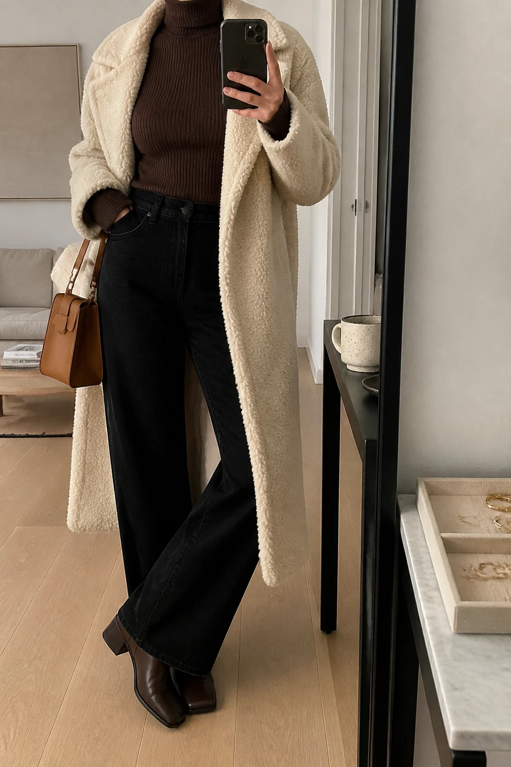 Image Prompt: Mirror selfie of a woman wearing a long cream oversized teddy-fleece coat over a chocolate brown fitted fine-ribbed turtleneck sweater, solid black high-waisted wide-leg denim jeans, dark brown leather square-toe ankle boots with low block heels, and a small structured caramel leather handbag, holding a phone in front of a mirror, cropped at the NECK so the face does not show, with a relaxed casual selfie pose, in a stylish city apartment, soft indoor lighting, a coffee mug on a side table and a jewelry tray on the vanity, realistic cream teddy-fleece texture, natural chocolate ribbed knit stitches, black denim draping, latest iPhone photo quality, no additional clothing or accessories.