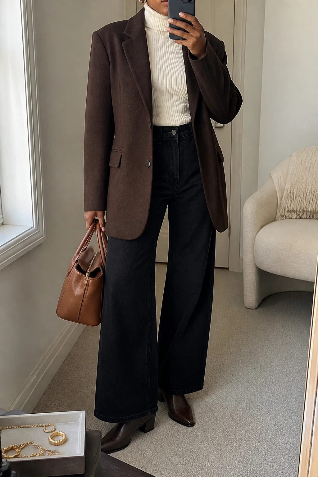 Image Prompt: Mirror selfie of a black woman wearing a chocolate brown oversized single-breasted wool-blend blazer over an ivory fitted ribbed turtleneck sweater, solid black high-waisted wide-leg denim jeans, dark brown leather pointed-toe ankle boots with low block heels, a structured caramel leather handbag, and delicate gold hoop earrings, holding a phone in front of a mirror, cropped at the NECK so the face does not show, with a relaxed casual selfie pose, in a minimal dressing room, soft natural window light, a jewelry tray on the vanity and a folded blanket on a chair, realistic chocolate wool blazer texture, natural ivory ribbed knit and black wide-leg denim folds, latest iPhone photo quality, no additional clothing or accessories.