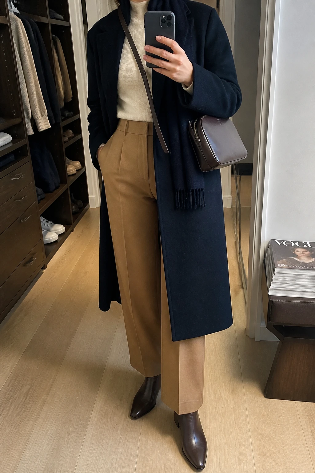 Image Prompt: Mirror selfie of a woman wearing a long navy single-breasted wool coat over an ivory fitted cashmere crewneck sweater, camel high-waisted straight-leg wool tailored trousers, chocolate brown waterproof leather pointed-toe ankle boots with low block heels and rubber soles, a structured dark brown leather zippered crossbody bag, and a navy cashmere scarf, holding a phone in front of a mirror, cropped at the NECK so the face does not show, with a relaxed casual selfie pose, in a walk-in closet mirror, soft indoor lighting, an open closet with clothes visible and a stack of fashion magazines on a side table, realistic navy wool fabric, natural camel trouser folds and elegant winter styling, latest iPhone photo quality, no additional clothing or accessories.