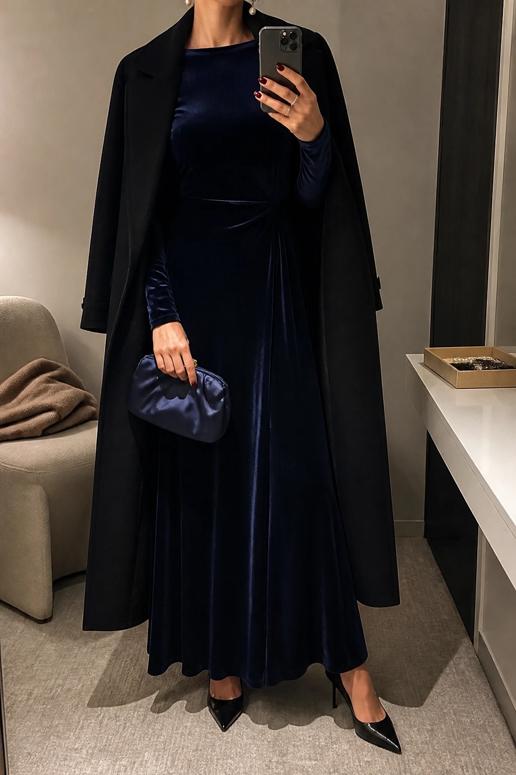 Image Prompt: Mirror selfie of a woman wearing a midnight navy long-sleeve velvet maxi dress with a fitted bodice and softly flared skirt, black pointed-toe suede heels, a small navy satin evening clutch, classic white pearl drop earrings, and a long black wool coat worn open, holding a phone in front of a mirror, cropped at the NECK so the face does not show, with a relaxed casual selfie pose, in a minimal dressing room, warm moody lighting, a folded blanket on a chair and a jewelry tray on the vanity, realistic navy velvet texture and natural long dress folds, latest iPhone photo quality, no additional clothing or accessories.