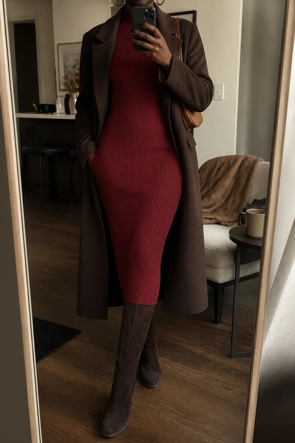 Image Prompt: Mirror selfie of a black woman wearing a deep red long-sleeve fitted ribbed knit midi dress with a high neckline and softly defined waist, chocolate brown suede knee-high boots with block heels, a long dark brown tailored wool coat worn open, a small caramel leather shoulder bag, and delicate gold hoop earrings, holding a phone in front of a full-length mirror, cropped at the NECK so the face does not show, with a relaxed casual selfie pose, in a stylish city apartment, warm moody lighting, a coffee mug on a side table and a folded blanket on a chair, realistic deep red knit texture, natural dark brown wool folds, subtle suede finish, latest iPhone photo quality, no additional clothing or accessories.