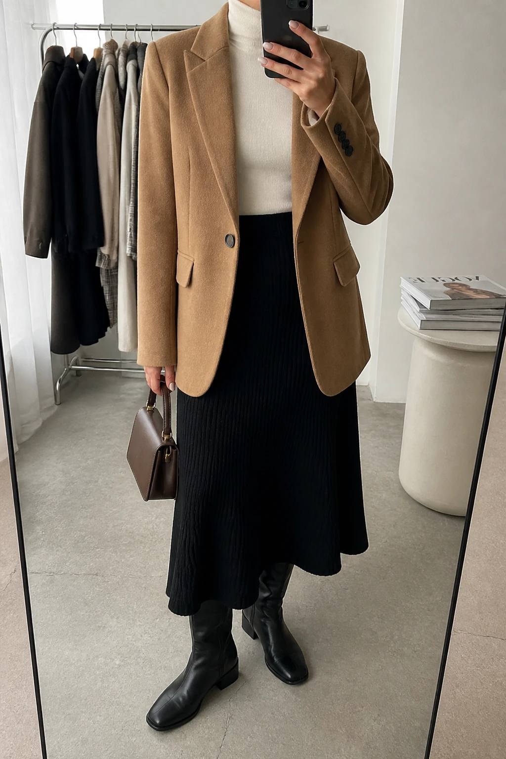 Image Prompt: Mirror selfie of a woman wearing a camel tailored wool-blend blazer over an ivory fine-knit fitted turtleneck sweater, a black ribbed knit A-line midi skirt, black leather knee-high low block-heel boots, a small structured chocolate brown leather handbag, and delicate gold stud earrings, holding a phone in front of a mirror, cropped at the NECK so the face does not show, with a relaxed casual selfie pose, in a fashion studio mirror corner, soft natural window light, a clothing rack with garments behind and a stack of fashion magazines on a side table, realistic camel wool texture and natural black knit skirt folds, latest iPhone photo quality, no additional clothing or accessories.