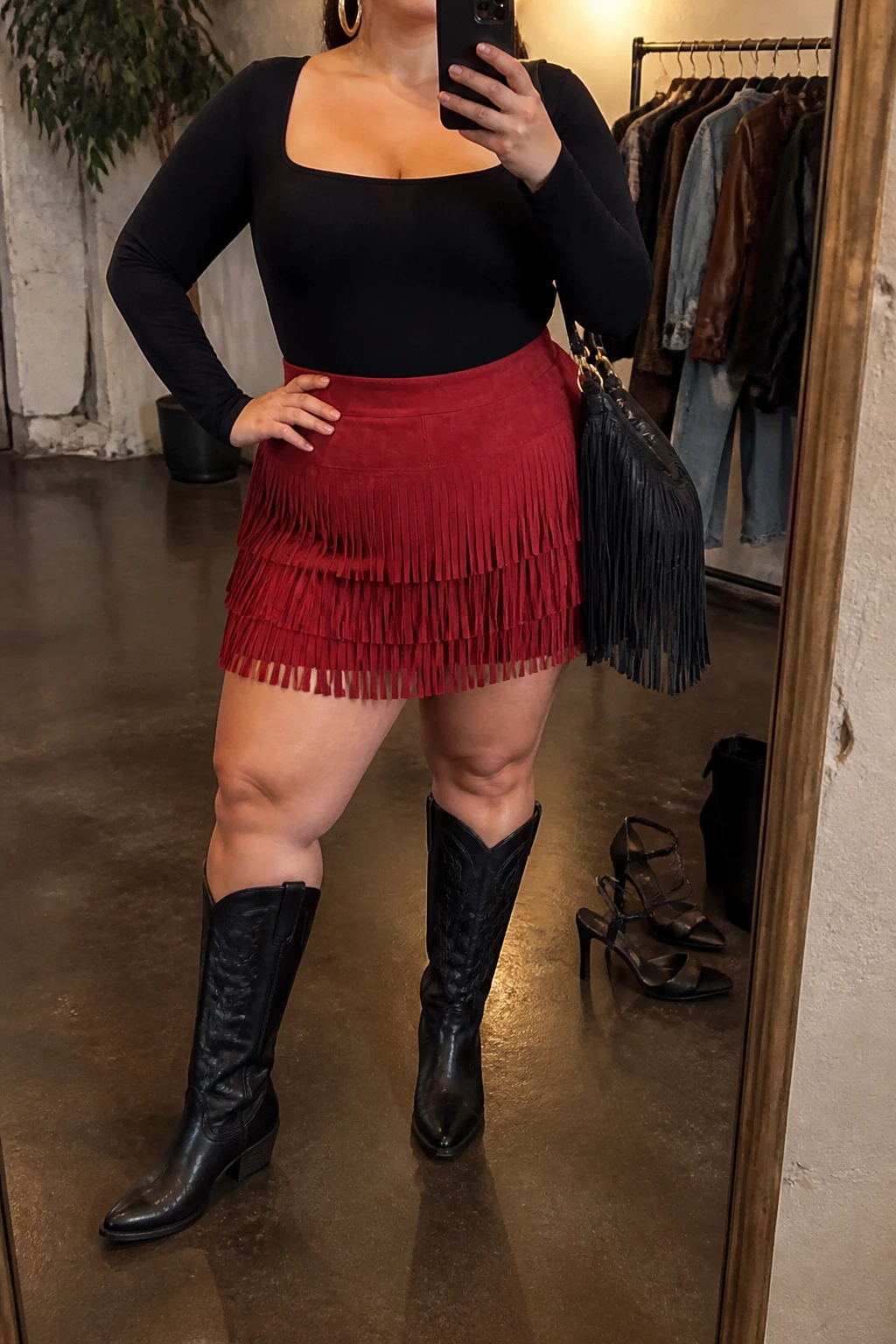 Mirror selfie of a plus size woman wearing a high-waisted red suede mini skirt with layered fringe, a fitted black long-sleeve square-neck bodysuit, black leather knee-high cowboy boots with low stacked heels, a small black fringe shoulder bag, and chunky gold hoop earrings, clearly showing the exact clothing items, red suede fringe, black fitted top, boots, bag, and accessories listed, holding a phone in front of a mirror, cropped at the neck so her face does not show, relaxed energetic selfie stance, in a fashion studio mirror corner with warm moody lighting, a clothing rack with garments behind her and a pair of heels on the floor nearby, realistic concert outfit, latest iPhone photo quality.