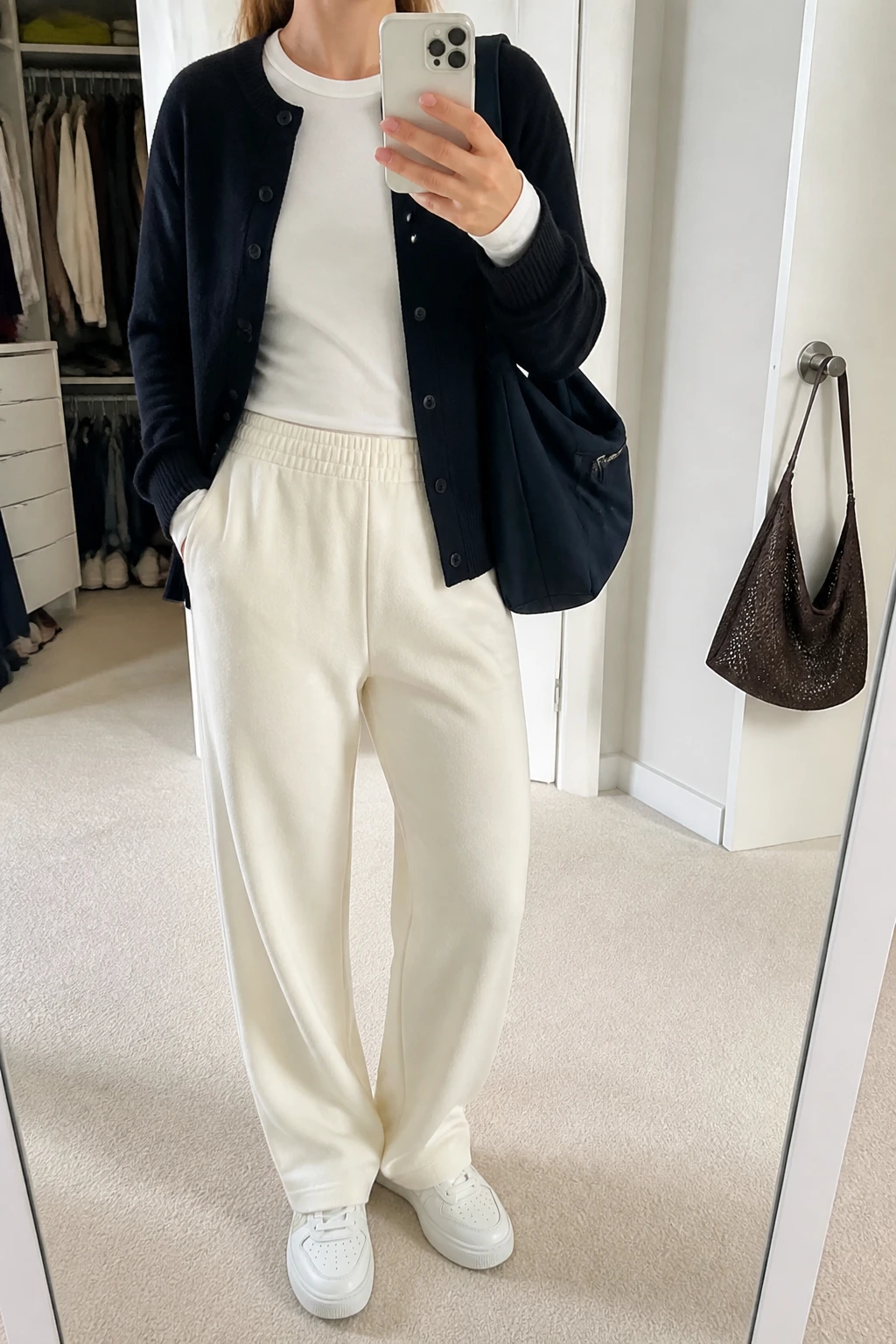 Image Prompt: Mirror selfie of a woman wearing a navy oversized fine-knit button-front cardigan over a white fitted cotton long-sleeve crewneck top, cream high-waisted relaxed straight-leg knit trousers with an elastic waistband, white cushioned leather sneakers, and a medium navy nylon zippered shoulder tote bag, holding a phone in front of a mirror, cropped at the NECK so the face does not show, with a relaxed casual selfie pose, in a walk-in closet mirror, soft natural window light, an open closet with clothes visible and a handbag hanging on a door handle, realistic navy cardigan texture, natural cream knit trouser folds and comfortable travel styling, latest iPhone photo quality, no additional clothing or accessories.