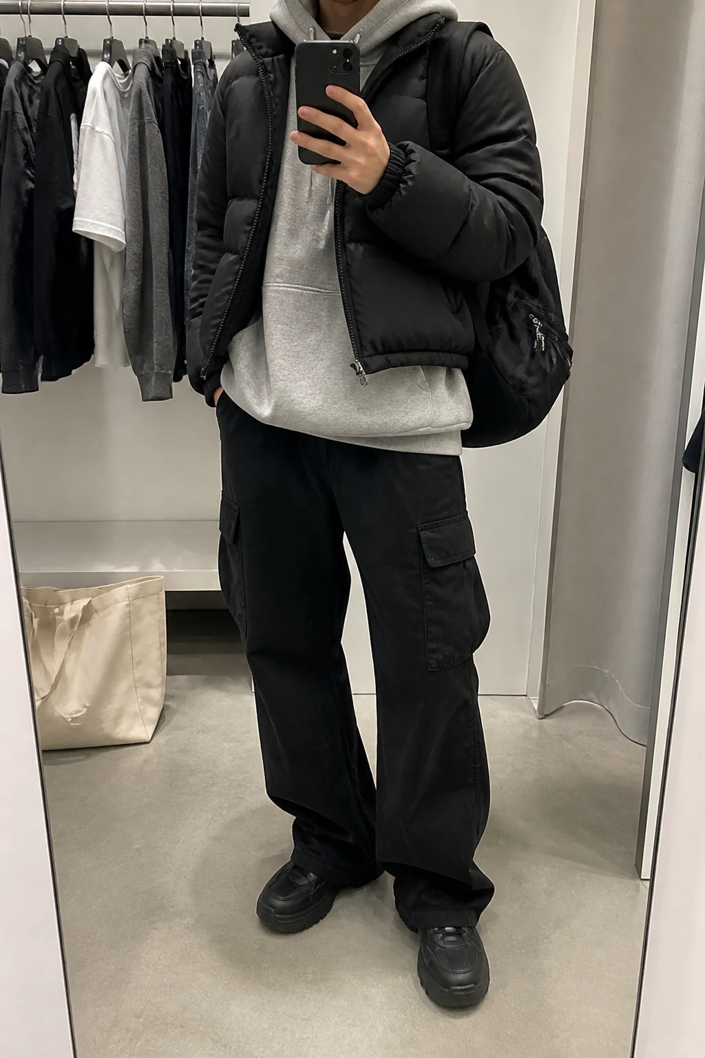 Image Prompt: Mirror selfie of a woman wearing a heather grey oversized fleece-lined pullover hoodie beneath a black cropped insulated puffer jacket, black baggy cotton-twill cargo trousers with large side pockets, black chunky leather sneakers with thick rubber soles, and a medium black nylon backpack with silver zippers, holding a phone in front of a mirror, cropped at the NECK so the face does not show, with a relaxed casual selfie pose, in a minimal dressing room, soft indoor lighting, a clothing rack with garments behind and a tote bag resting on the floor, realistic grey fleece texture, natural cargo trouser folds and slightly imperfect casual campus styling, latest iPhone photo quality, no additional clothing or accessories.