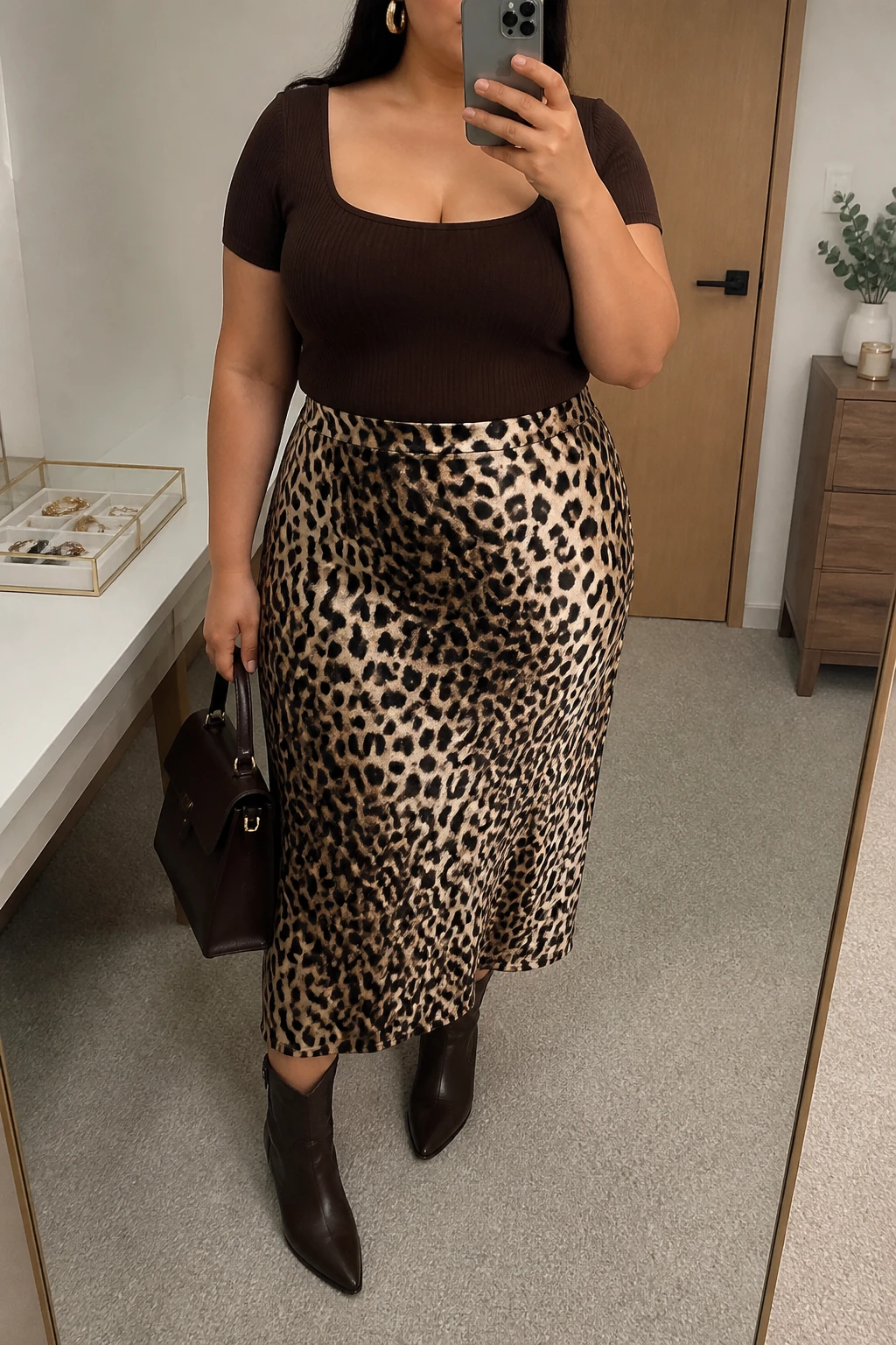 Mirror selfie of a plus size woman wearing a leopard print satin midi skirt with a soft bias-cut shape, a fitted chocolate brown short-sleeve square-neck knit top, chocolate brown pointed ankle cowboy boots, a structured dark brown handbag, and hammered gold hoop earrings, clearly showing the exact clothing items, leopard print, chocolate knit, brown boots, bag, and accessories listed, holding a phone in front of a mirror, cropped at the neck so her face is hidden, relaxed polished selfie stance, in a minimal dressing room with soft indoor lighting, a jewelry tray on the vanity and a small plant on a dresser, realistic dinner outfit, latest iPhone photo quality.