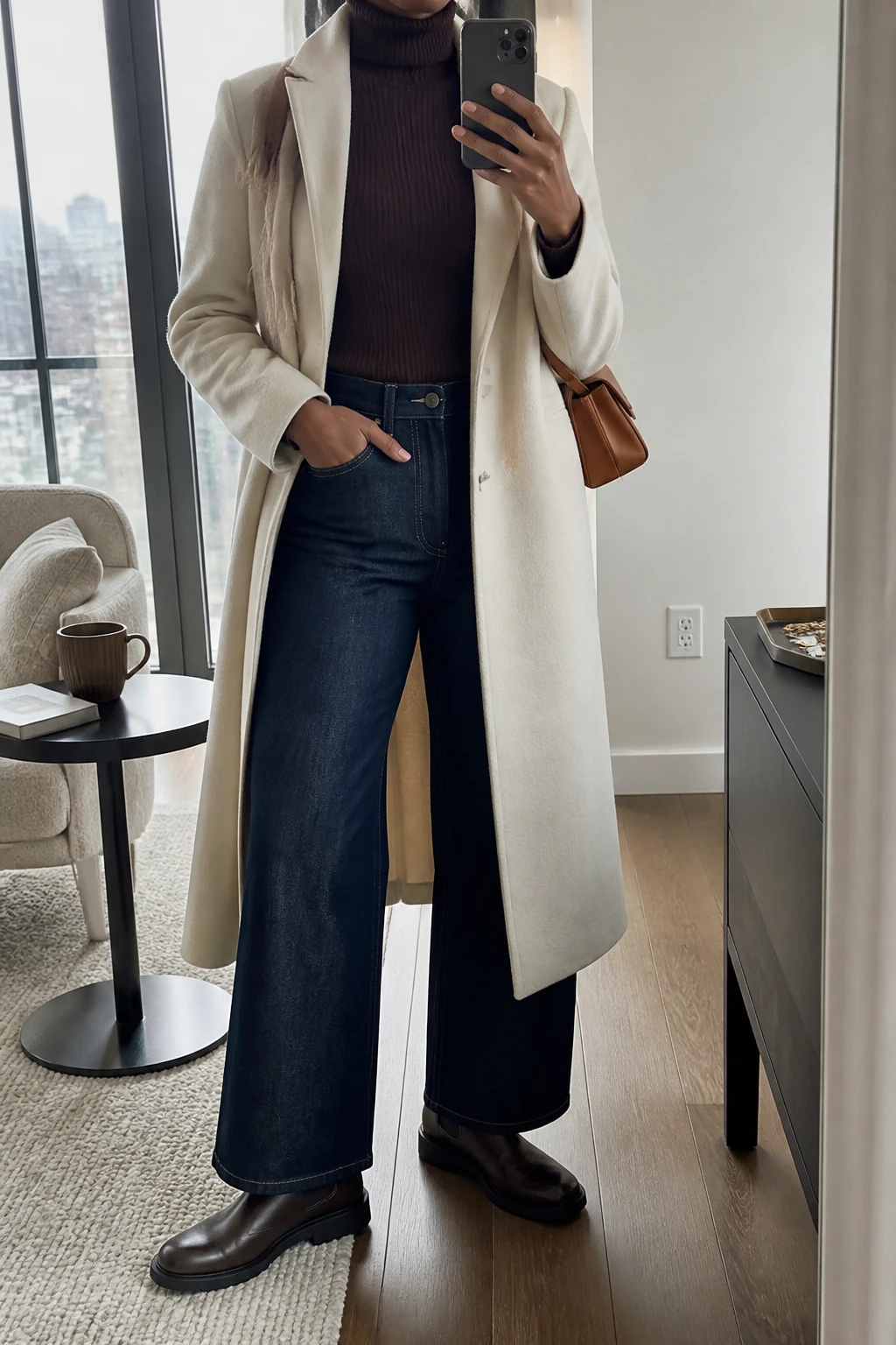 Image Prompt: Mirror selfie of a black woman wearing a long cream single-breasted tailored wool coat over a chocolate brown fitted ribbed turtleneck sweater, dark indigo high-waisted wide-leg denim jeans, dark brown leather flat Chelsea boots with rubber soles, a small structured caramel leather shoulder bag, and a beige cashmere scarf, holding a phone in front of a mirror, cropped at the NECK so the face does not show, with a relaxed casual selfie pose, in a stylish city apartment, soft natural window light, a coffee mug on a side table and a jewelry tray on the vanity, realistic cream wool coat texture, natural dark denim folds, chocolate ribbed knit fabric, latest iPhone photo quality, no additional clothing or accessories.