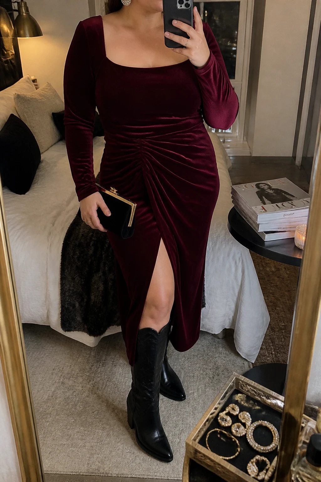 Mirror selfie of a plus size woman wearing a burgundy velvet midi dress with long sleeves, a square neckline, soft waist gathering, and a front slit, black leather knee-high cowboy boots with low stacked heels, a small black velvet clutch, and silver crystal stud earrings, clearly showing the exact dress, burgundy velvet, waist gathering, black boots, clutch, and accessories listed, holding a phone in front of a mirror, cropped at the neck so the face is hidden, relaxed elegant selfie pose, in a cozy bedroom mirror setup with warm moody lighting, a stack of fashion magazines on a side table and a jewelry tray on the vanity, realistic winter evening styling, latest iPhone photo quality.