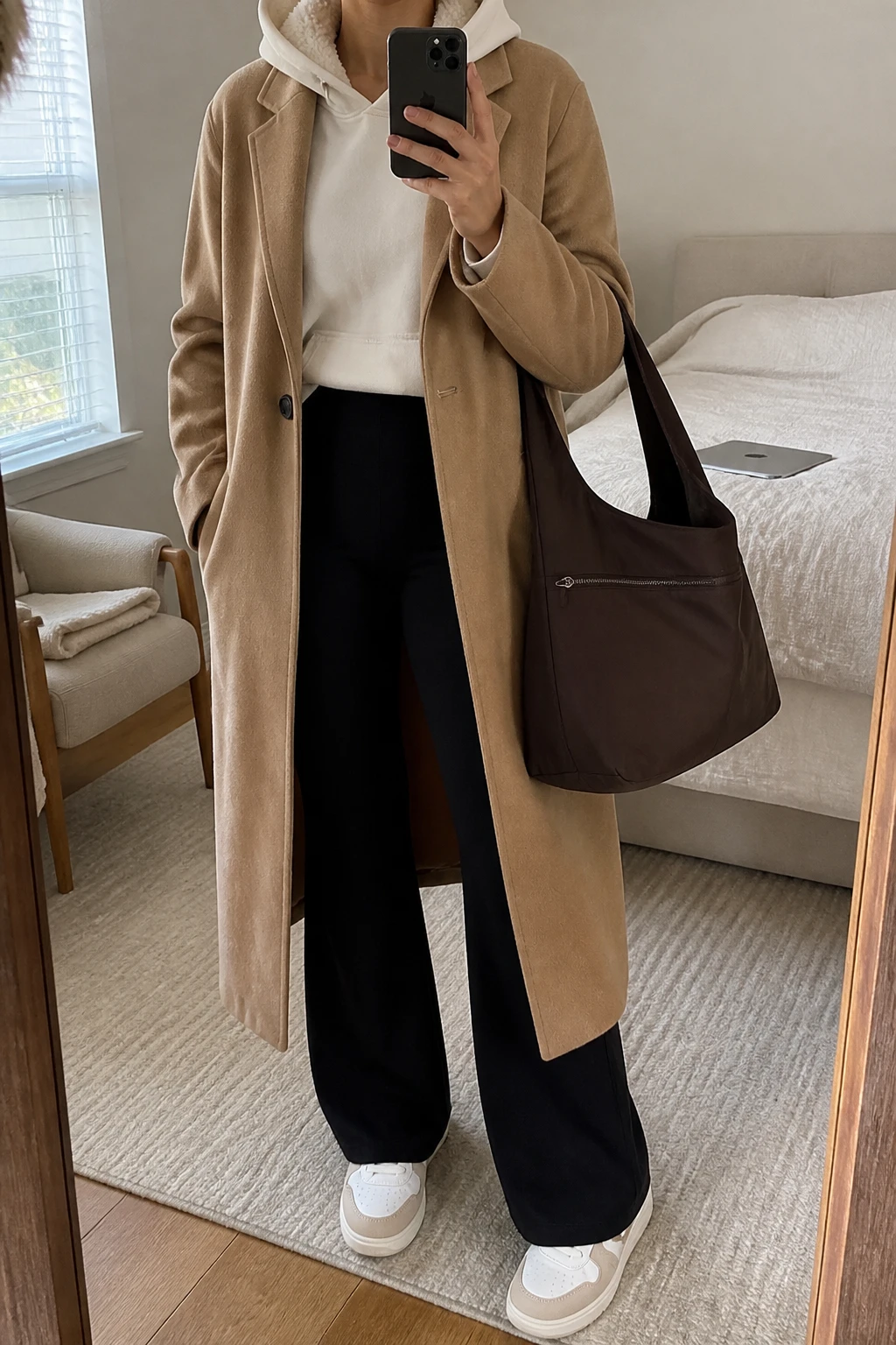 Image Prompt: Mirror selfie of a woman wearing a long camel single-breasted wool-blend coat left open over an oversized cream fleece-lined hoodie, black high-waisted thick stretch wide-leg leggings, white cushioned leather sneakers with beige detailing, and a large chocolate brown zippered nylon tote bag with comfortable shoulder straps, holding a phone in front of a mirror, cropped at the NECK so the face does not show, with a relaxed casual selfie pose, in a cozy bedroom mirror setup, soft natural window light, a folded blanket on a chair and a laptop on the bed, realistic camel wool texture, natural fleece hoodie folds and comfortable travel proportions, latest iPhone photo quality, no additional clothing or accessories.