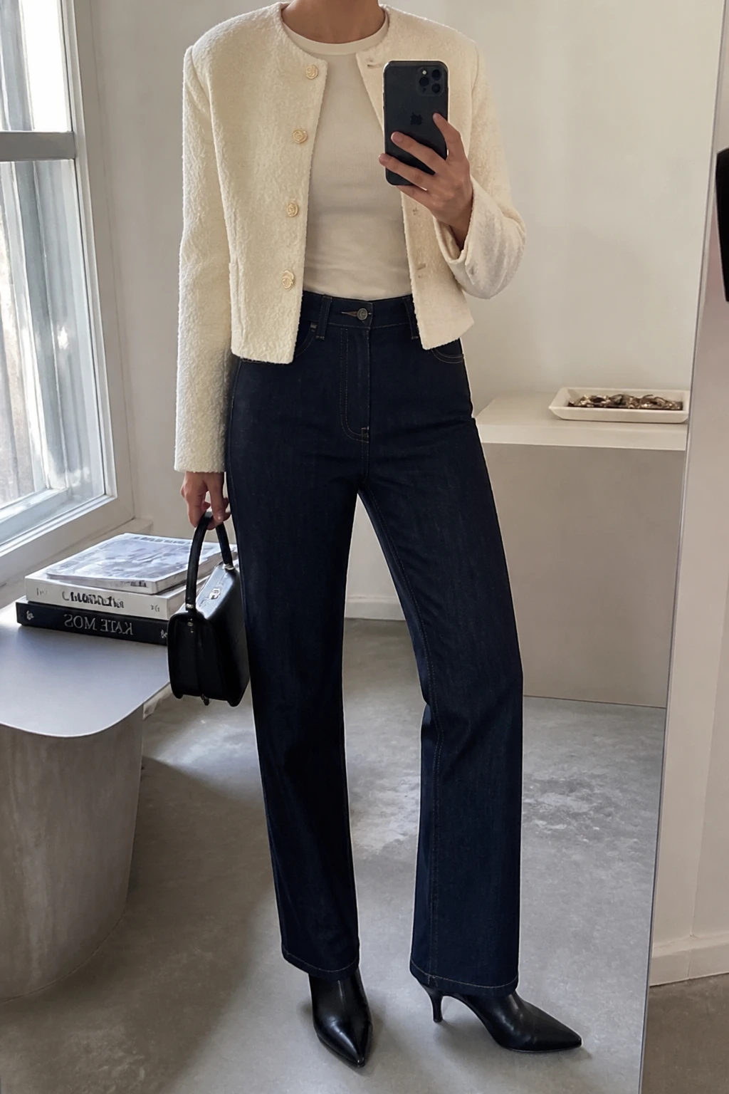 Image Prompt: Mirror selfie of a woman wearing an ivory cropped bouclé jacket with gold buttons, left open over a cream fitted fine-knit crewneck top, dark indigo high-waisted straight-leg jeans, black leather pointed-toe heeled ankle boots, and carrying a small structured black leather top-handle bag, holding a phone in front of a mirror, cropped at the NECK so the face does not show, with a relaxed casual selfie pose, in a fashion studio mirror corner, soft natural window light, a stack of fashion magazines on a side table and a jewelry tray on the vanity, believable bouclé texture, subtle denim creases, natural unposed appearance, latest iPhone photo quality, no additional clothing or accessories.