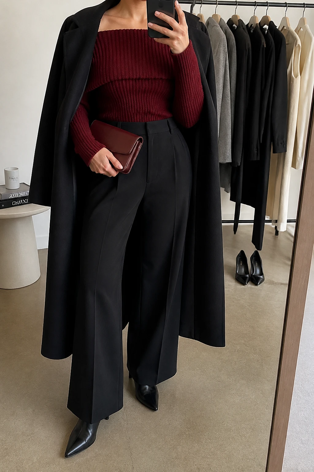 Image Prompt: Mirror selfie of a woman wearing a burgundy fitted off-shoulder chunky-knit sweater with a folded neckline, black high-waisted wide-leg tailored wool trousers, black leather pointed-toe heeled ankle boots, a long black wool coat draped over the shoulders, and a small burgundy leather clutch, holding a phone in front of a mirror, cropped at the NECK so the face does not show, with a relaxed casual selfie pose, in a fashion studio mirror corner, warm soft indoor lighting, a clothing rack with garments behind and a pair of heels on the floor nearby, realistic burgundy knit texture and natural tailored trouser folds, latest iPhone photo quality, no additional clothing or accessories.