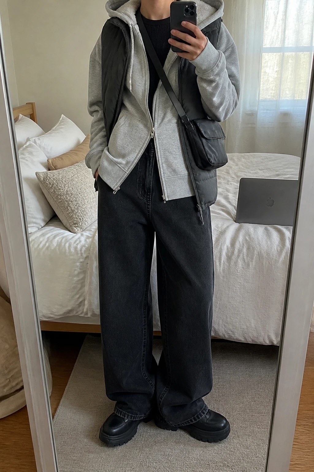 Image Prompt: Mirror selfie of a woman wearing a heather grey oversized fleece-lined zip-up hoodie partly unzipped over a black fitted thermal crewneck top, washed black wide-leg denim jeans, a charcoal grey quilted sleeveless puffer vest worn open, black chunky platform sneakers with thick rubber soles, and a small black nylon crossbody bag, holding a phone in front of a mirror, cropped at the NECK so the face does not show, with a relaxed casual selfie pose, in a cozy bedroom mirror setup, natural window light, soft pillows on an unmade bed and a laptop on the bed, realistic grey fleece texture, natural baggy denim creases, latest iPhone photo quality, no additional clothing or accessories.