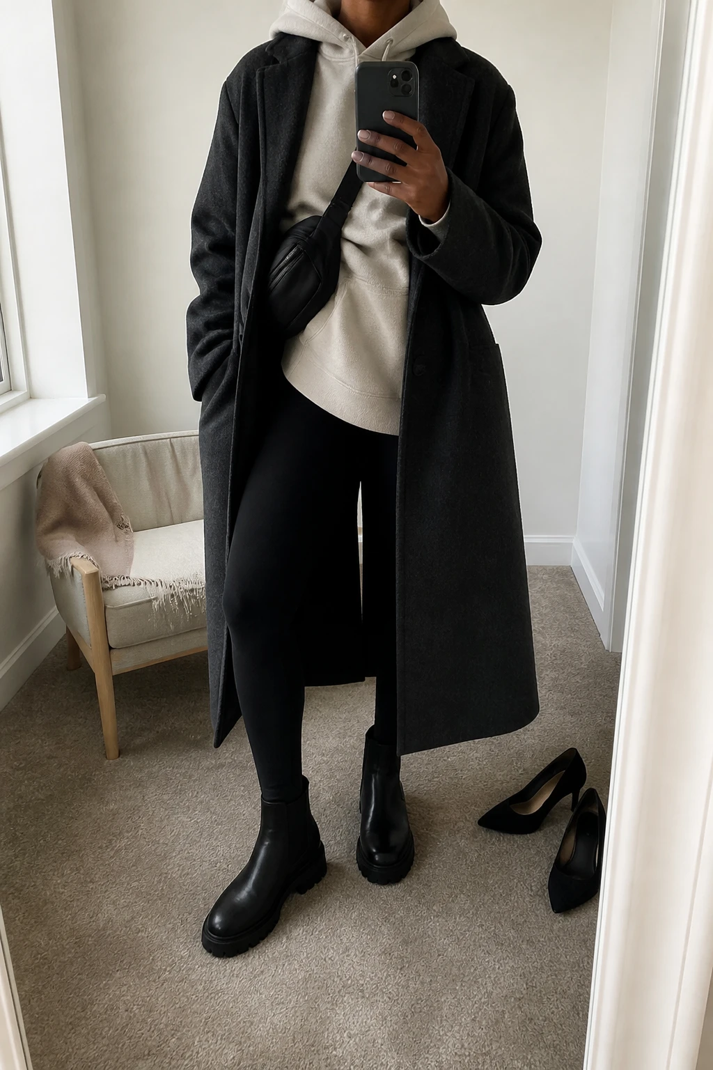 Image Prompt: Mirror selfie of a black woman wearing a long charcoal grey single-breasted wool coat left open over an oversized oatmeal fleece-lined hoodie, black thick thermal leggings, black leather chunky Chelsea boots with flat lug soles, and a black nylon crossbody bag, holding a phone in front of a mirror, cropped at the NECK so the face does not show, with a relaxed casual selfie pose, in a minimal dressing room, soft natural window light, a folded blanket on a chair and a pair of heels on the floor nearby, natural hoodie folds, realistic coat drape and casual winter layering, latest iPhone photo quality, no additional clothing or accessories.