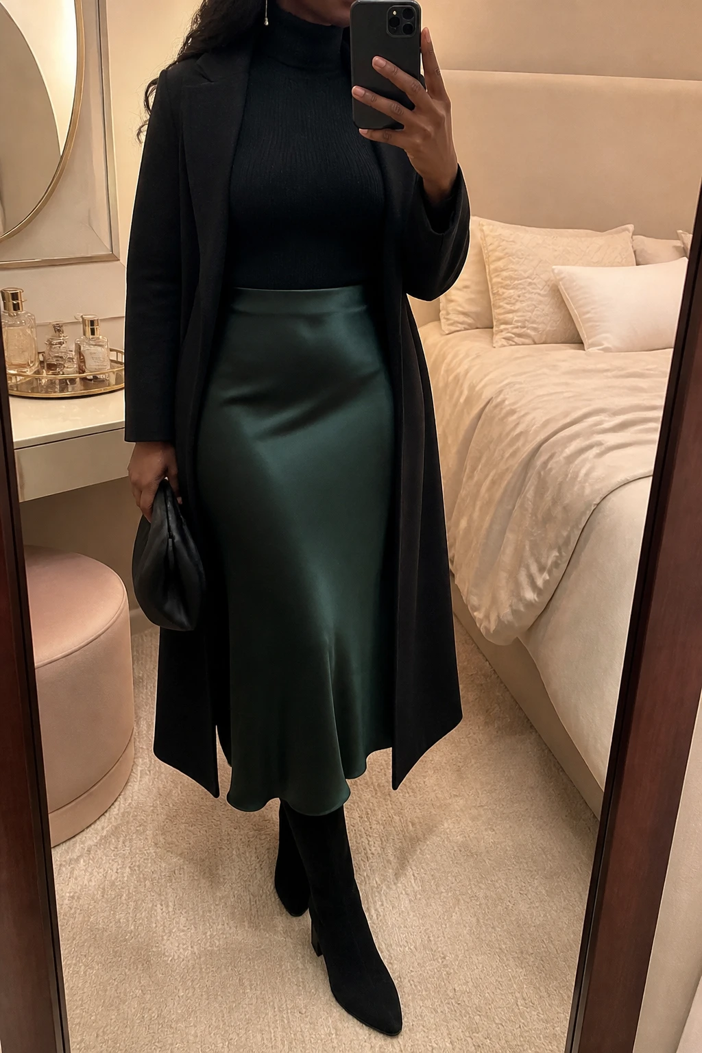 Image Prompt: Mirror selfie of a black woman wearing a black fitted cashmere turtleneck sweater tucked into a deep emerald green satin bias-cut midi skirt, black suede knee-high boots with low block heels, a long black tailored wool coat worn open, a small black satin clutch, and delicate silver drop earrings, holding a phone in front of a mirror, cropped at the NECK so the face does not show, with a relaxed casual selfie pose, in a feminine-themed bedroom, warm soft indoor lighting, a jewelry tray on the vanity and soft pillows on an unmade bed, realistic emerald green satin drape with gentle natural reflections, soft black knit texture, latest iPhone photo quality, no additional clothing or accessories.