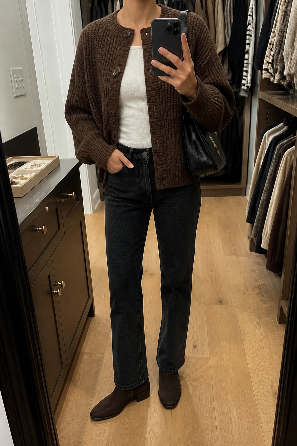 Image Prompt: Mirror selfie of a woman wearing a chocolate brown chunky-knit button-front cardigan over an ivory fitted ribbed cotton top, washed black straight-leg jeans, dark brown suede low-heeled ankle boots, and a small black leather shoulder bag, holding a phone in front of a mirror, cropped at the NECK so the face does not show, with a relaxed casual selfie pose, in a walk-in closet mirror, warm soft indoor lighting, an open closet with clothes visible and a jewelry tray on the vanity, true-to-life brown knit texture, natural denim creases, latest iPhone photo quality, no additional clothing or accessories.