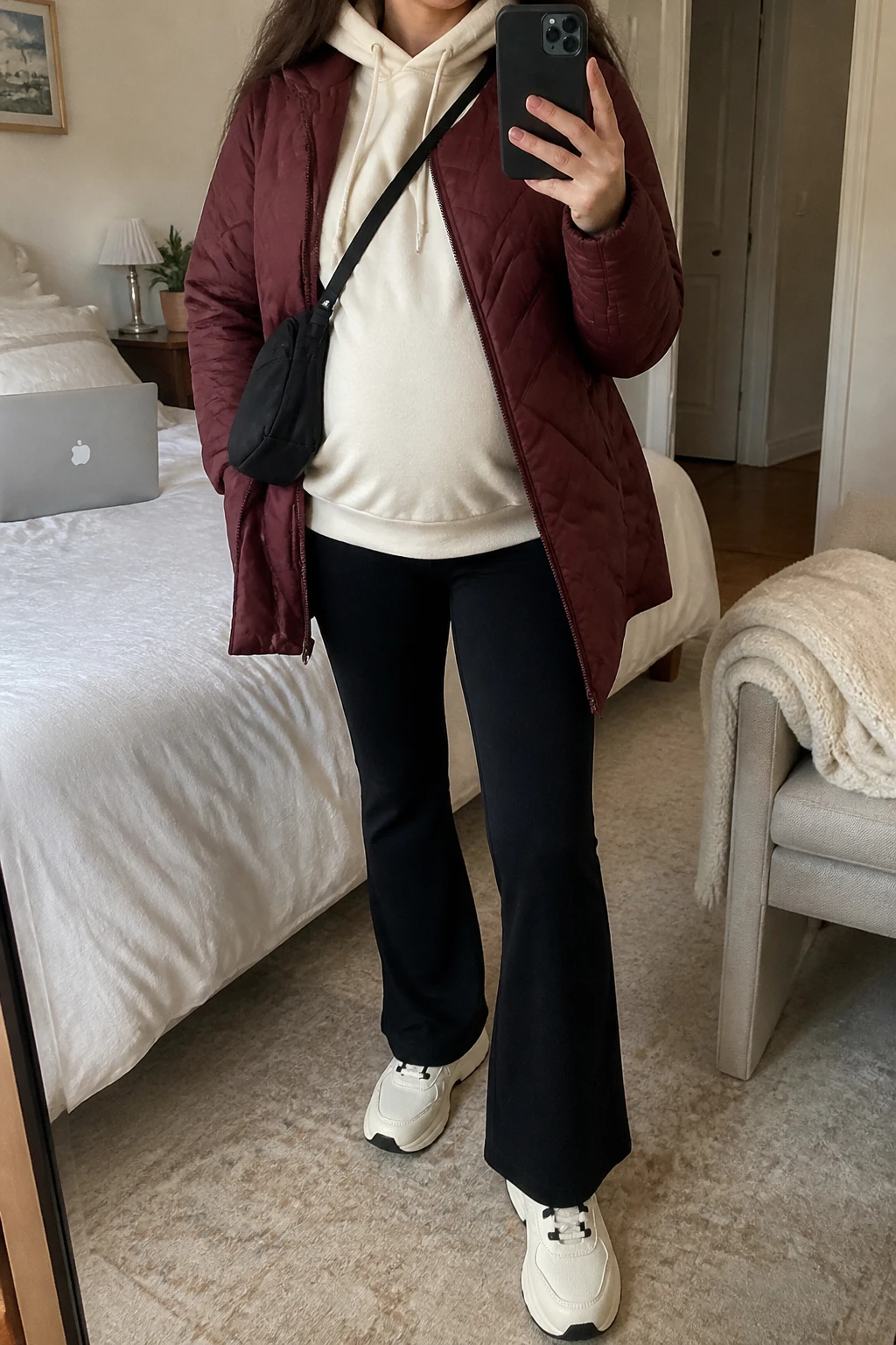 Image Prompt: Mirror selfie of a visibly pregnant woman wearing a burgundy lightweight insulated quilted hip-length jacket left open over an ivory oversized fleece-lined maternity hoodie with comfortable space around her baby bump, black high-waisted over-the-bump fleece-lined flared maternity leggings, cream cushioned chunky leather sneakers with black detailing, and a small black nylon crossbody bag, holding a phone in front of a mirror, cropped at the NECK so the face does not show, with a relaxed casual selfie pose and naturally visible pregnancy shape, in a cozy bedroom mirror setup, soft indoor lighting, a laptop on the bed and a folded blanket on a chair, realistic burgundy quilted texture, natural ivory fleece folds, comfortable maternity silhouette, latest iPhone photo quality, no additional clothing or accessories.